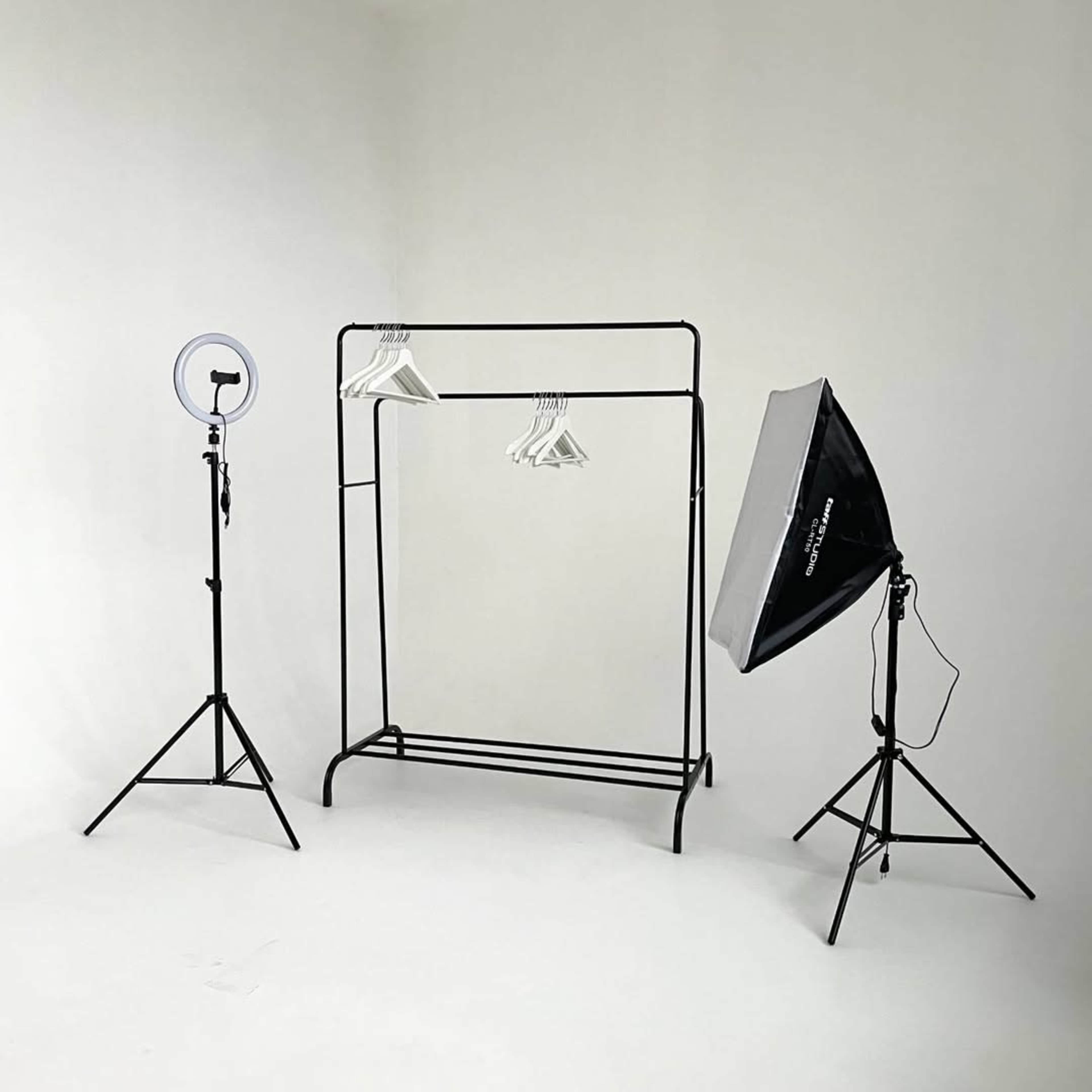 A minimalist photography setup features a black clothing rack with several hangers, a ring light on a stand, and an umbrella light on another stand, all set against a plain white backdrop.