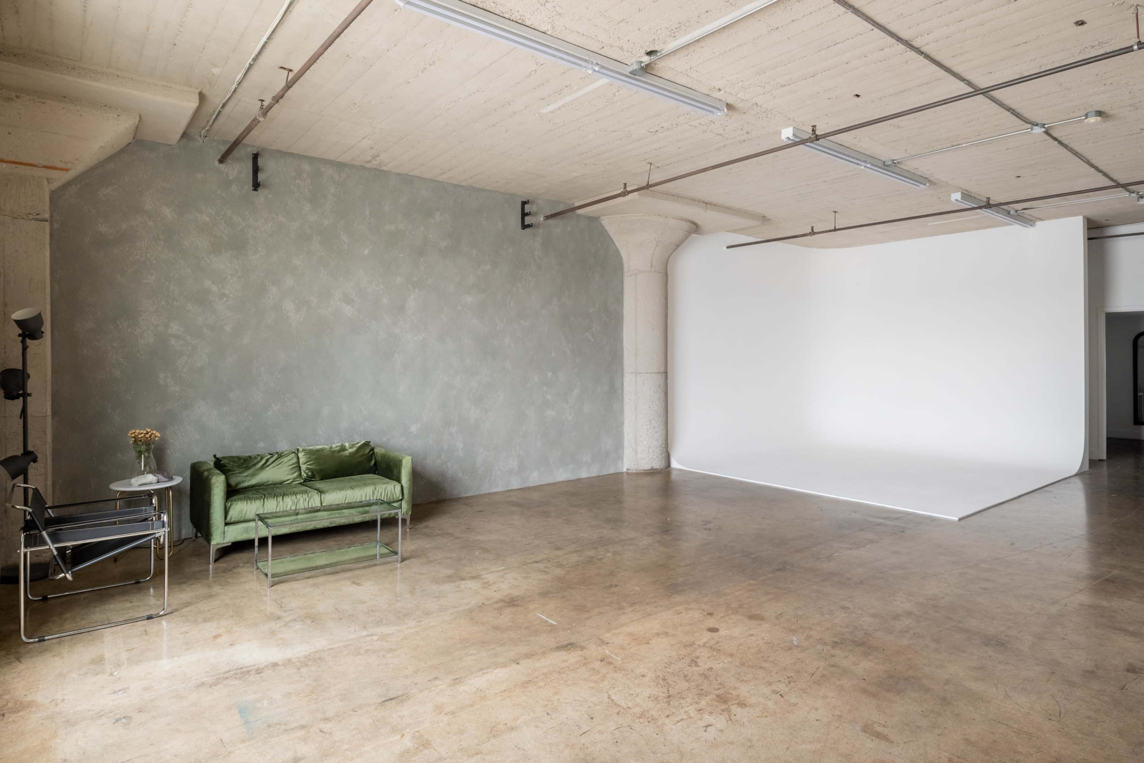 The image shows a spacious, minimalist studio featuring a green couch, a chair, and a blank white backdrop.