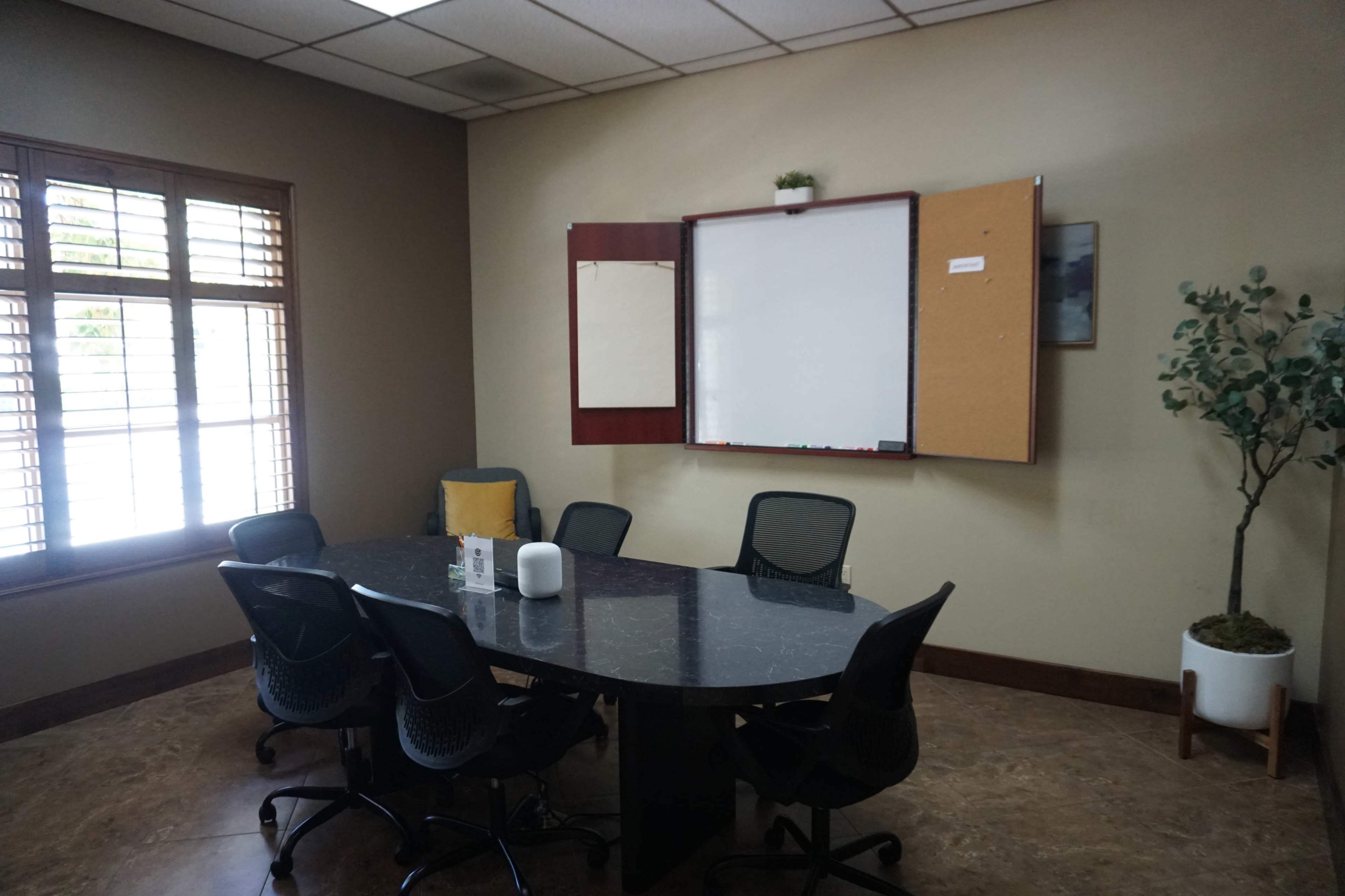 A conference room features a circular table surrounded by six chairs, with a whiteboard and bulletin board mounted on the wall.