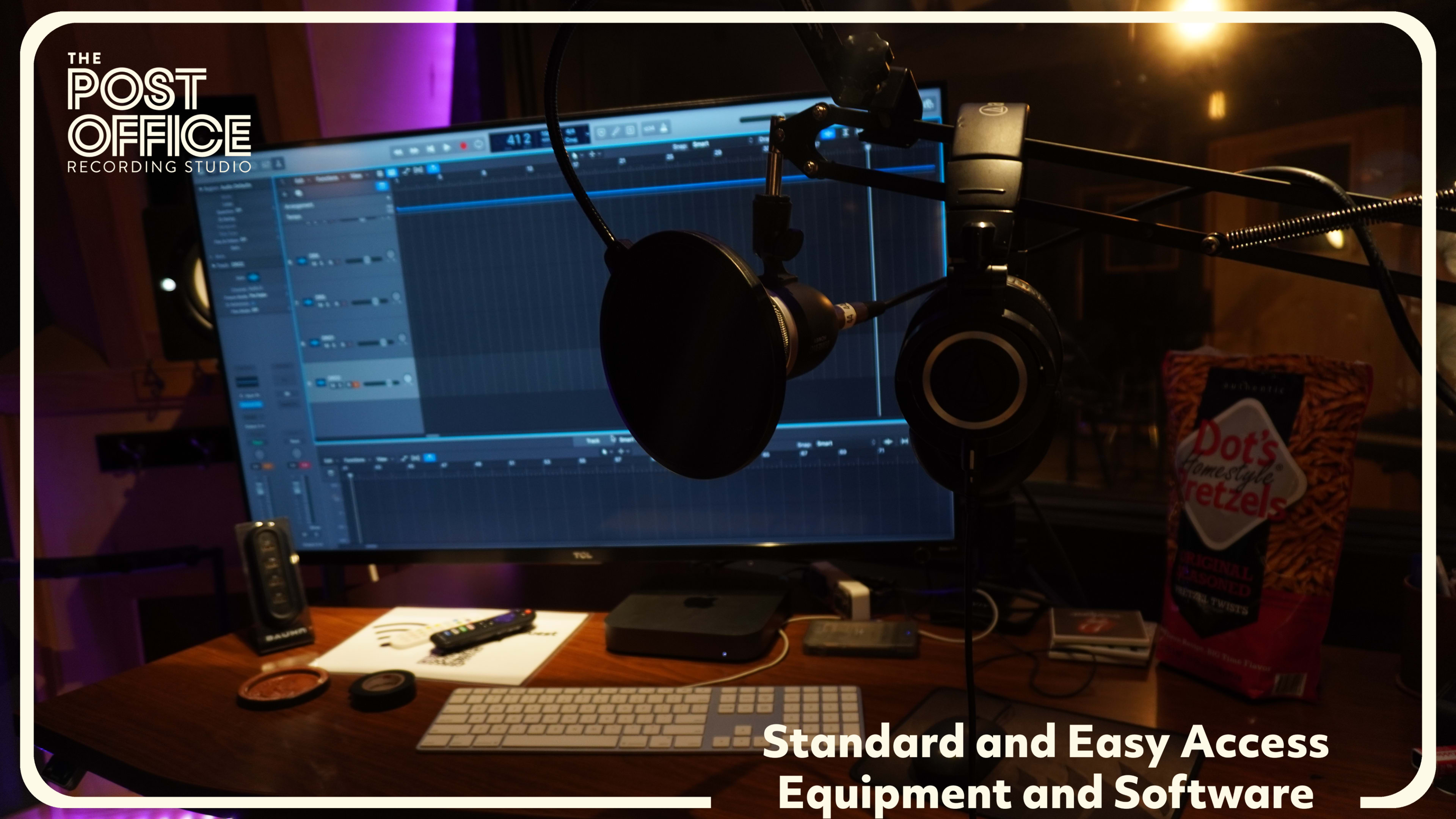A microphone and headphones hang in front of a computer monitor displaying audio editing software in a recording studio.
