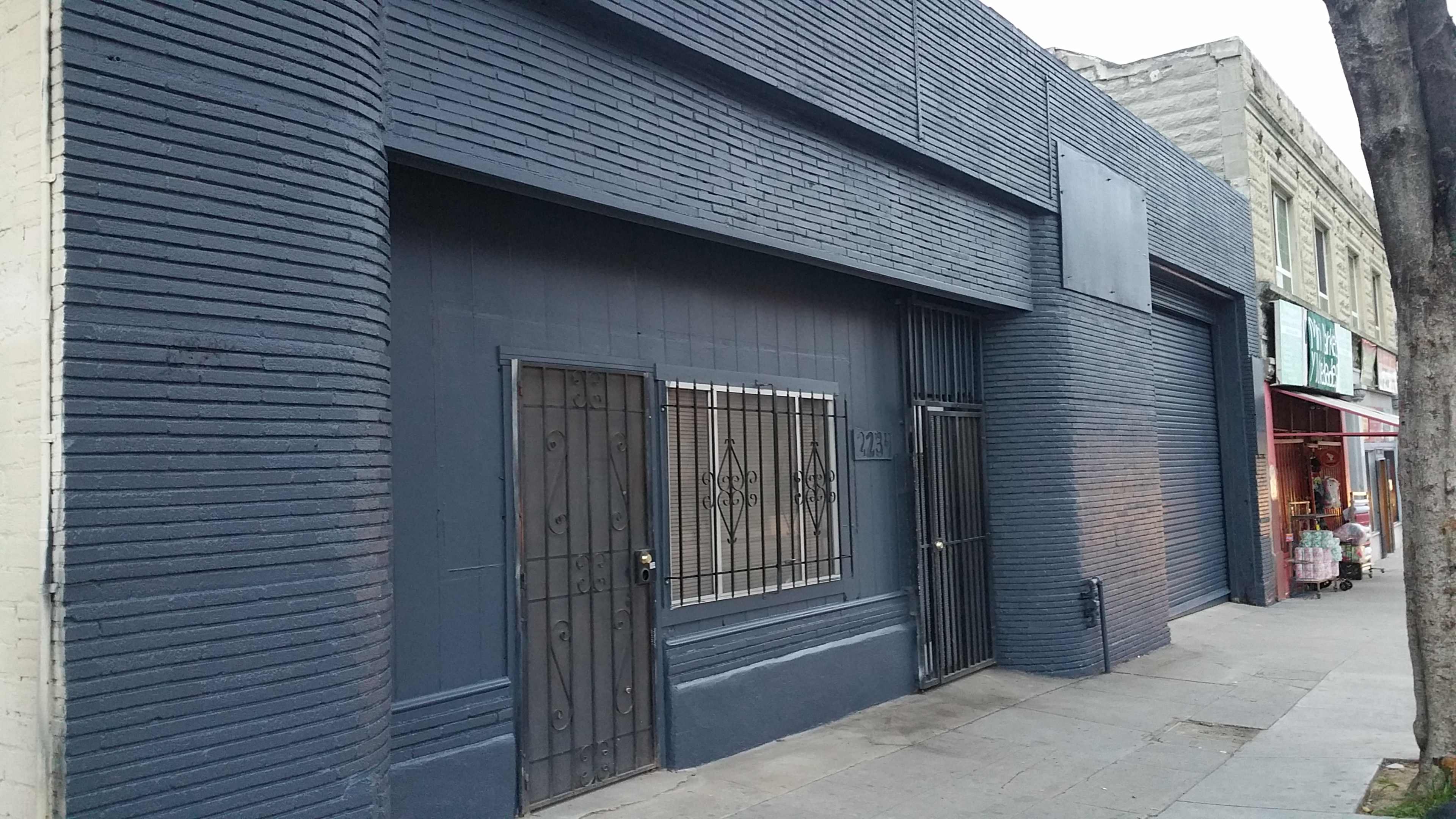 A gray-painted building with barred windows and a blank signboard stands along a sidewalk in an urban area.