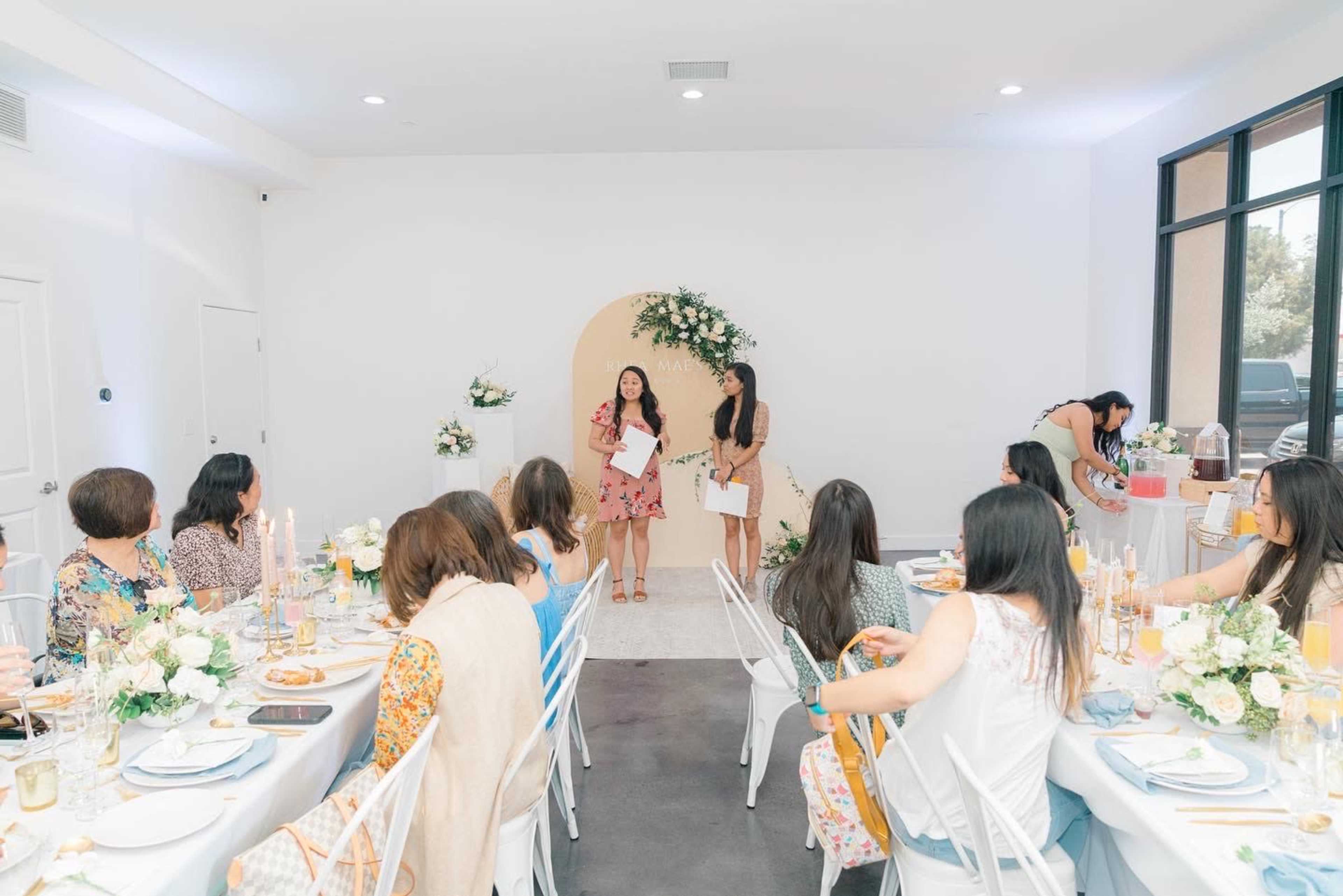 A group of women sit at elegantly set tables while two speakers present in front of a floral backdrop in a bright, modern event space.