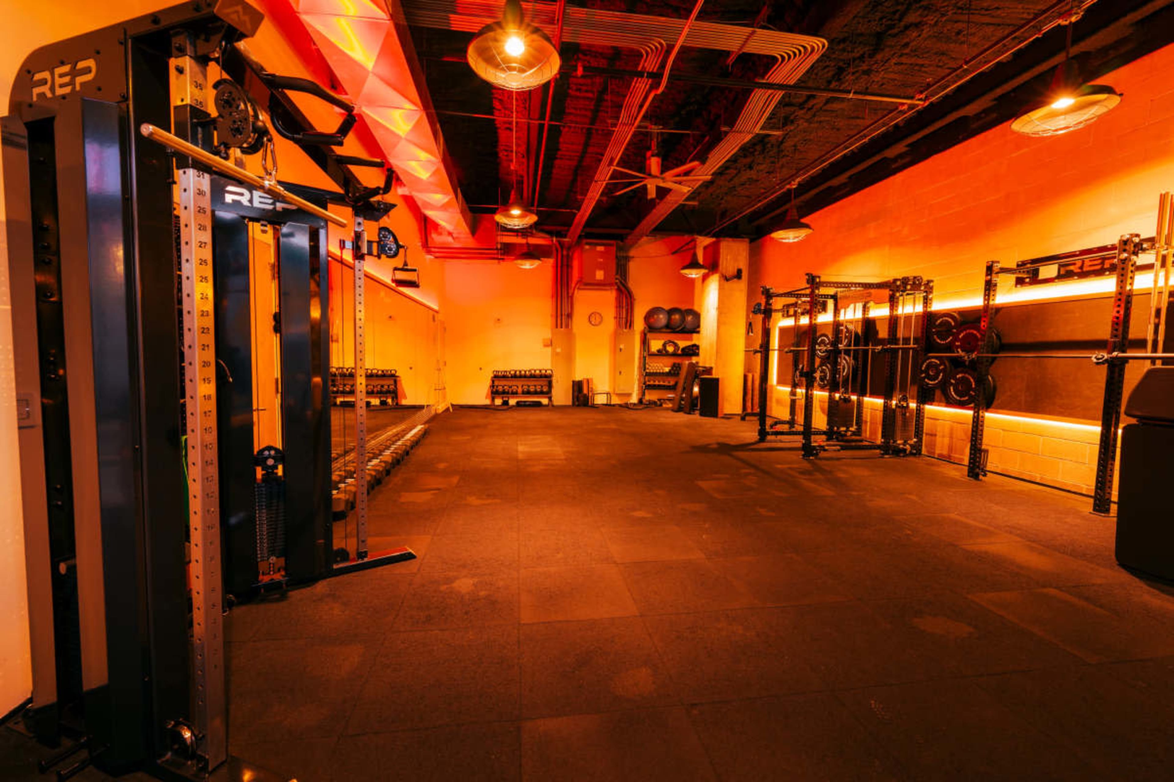 The image shows a well-equipped gym with multiple weightlifting stations and dim, amber lighting.
