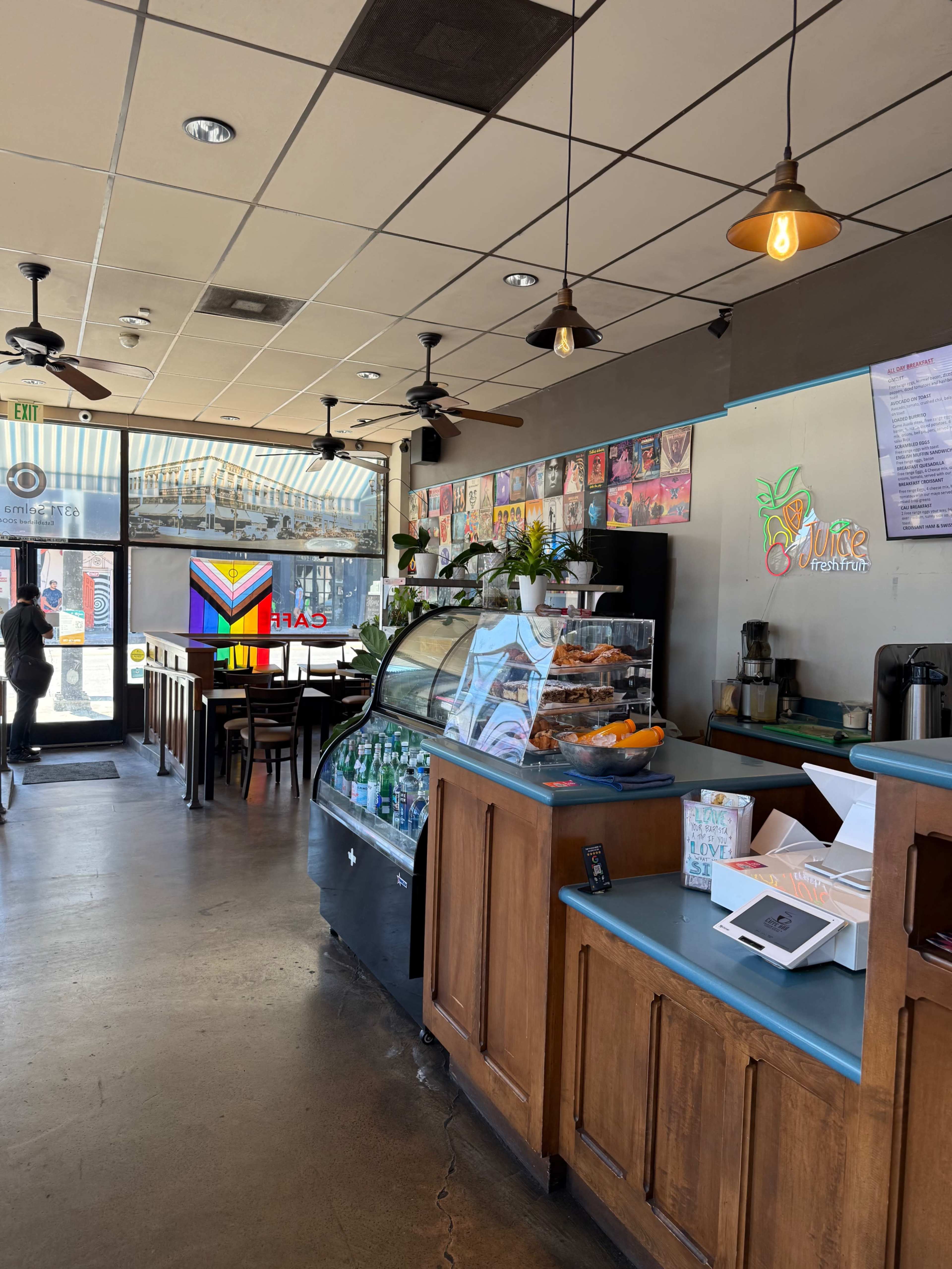 The image shows the interior of a café with a counter displaying baked goods and beverages, surrounded by tables and chairs, and large windows letting in natural light.