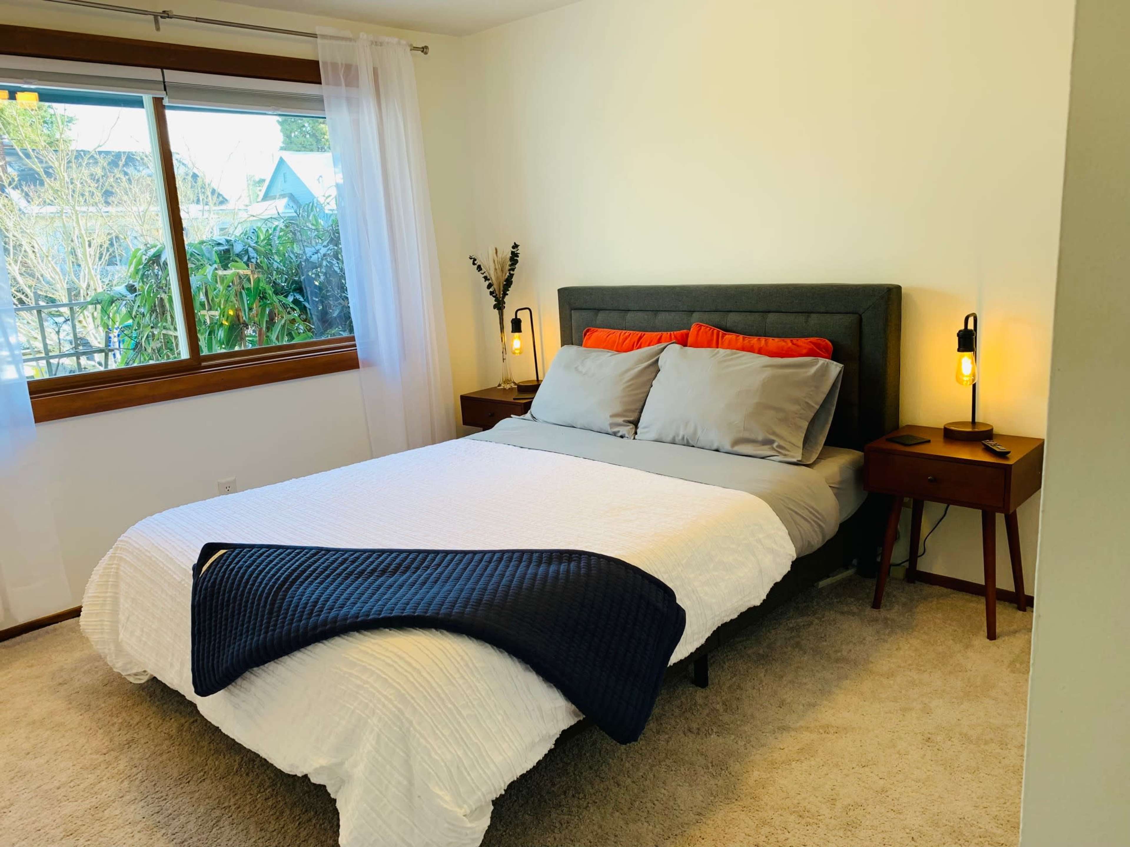 A neatly arranged bedroom features a gray upholstered bed with colorful pillows, flanked by wooden nightstands with lamps, and a large window letting in natural light.