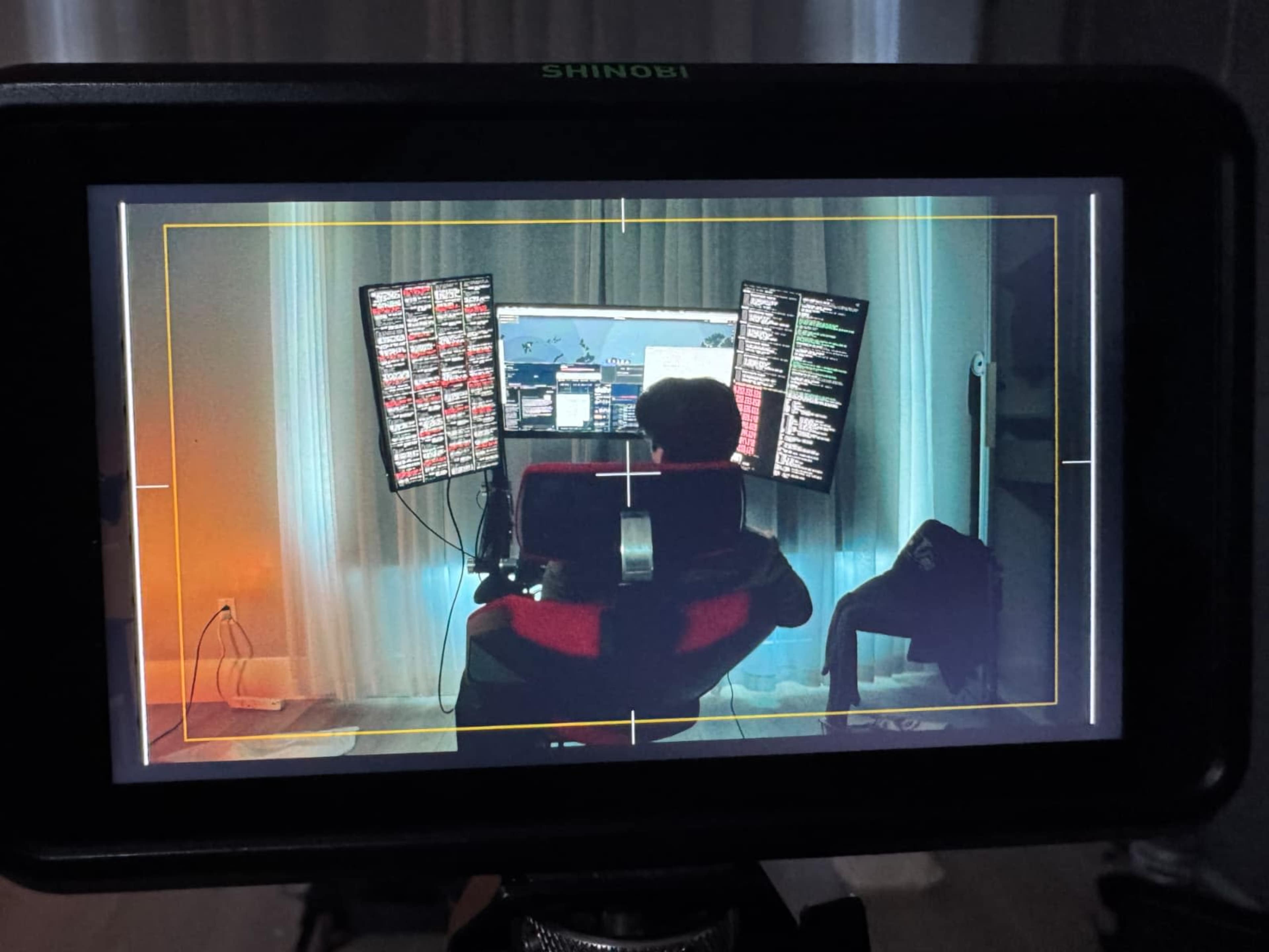 A person sits in a chair in front of a multi-monitor setup displaying data or code, with curtains in the background.