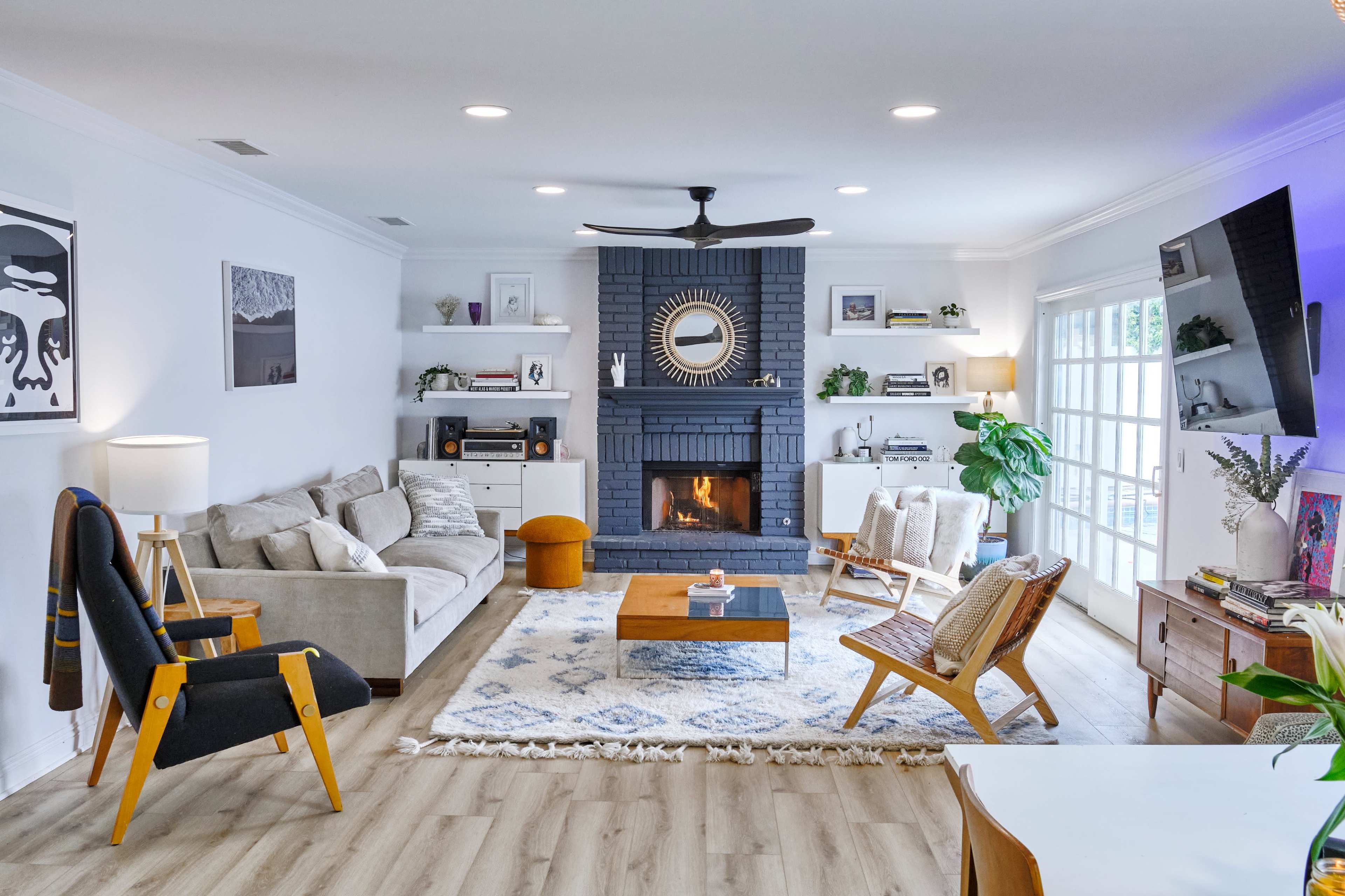 The living room features a modern layout with a gray brick fireplace, white walls, a large rug, and a combination of contemporary furniture arranged around a central coffee table.