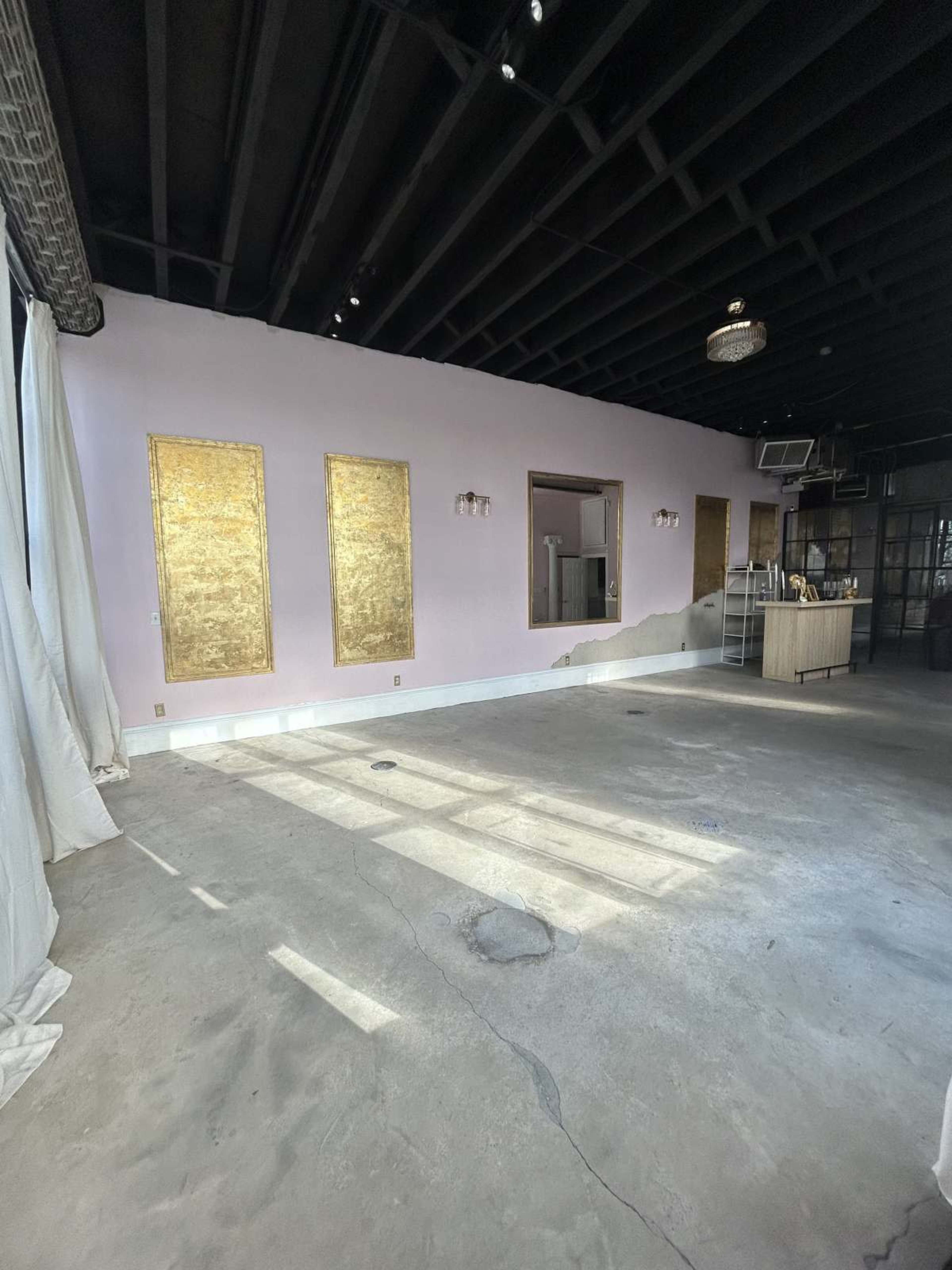 The image shows a spacious, partially furnished room with light pink walls and two gold-framed wall panels, featuring a concrete floor and large windows allowing natural light to enter.