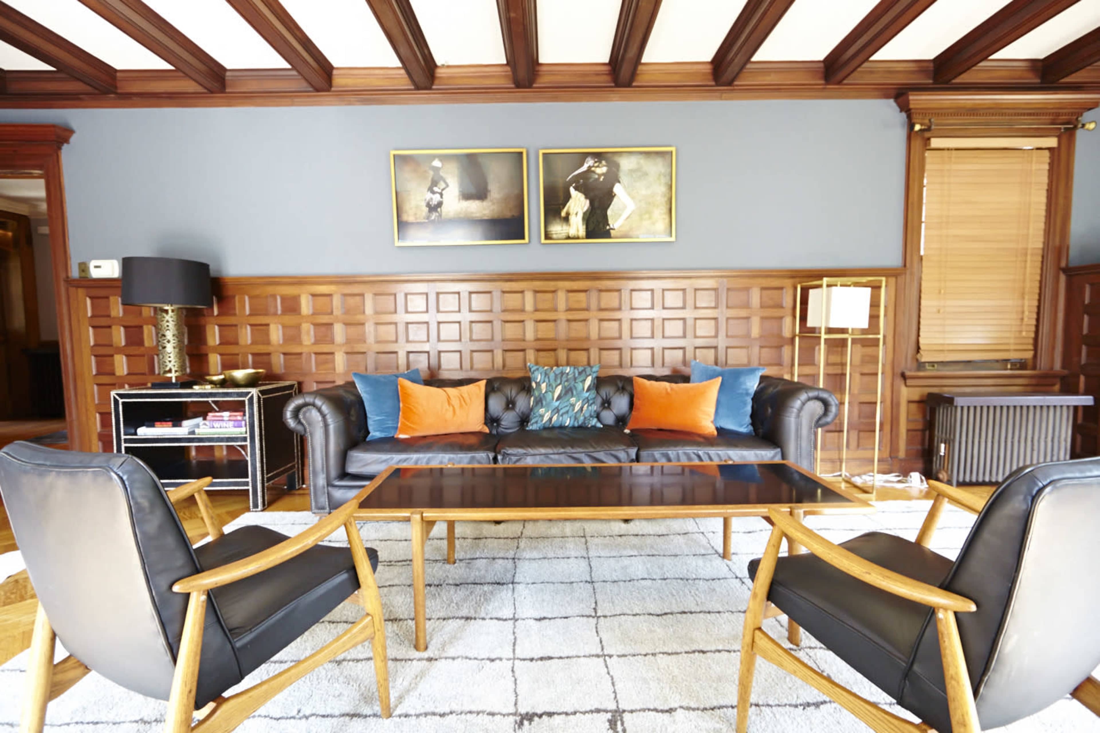 A living room features a dark leather sofa with colorful cushions, two mid-century chairs, a wooden coffee table, and framed artwork on the wall.