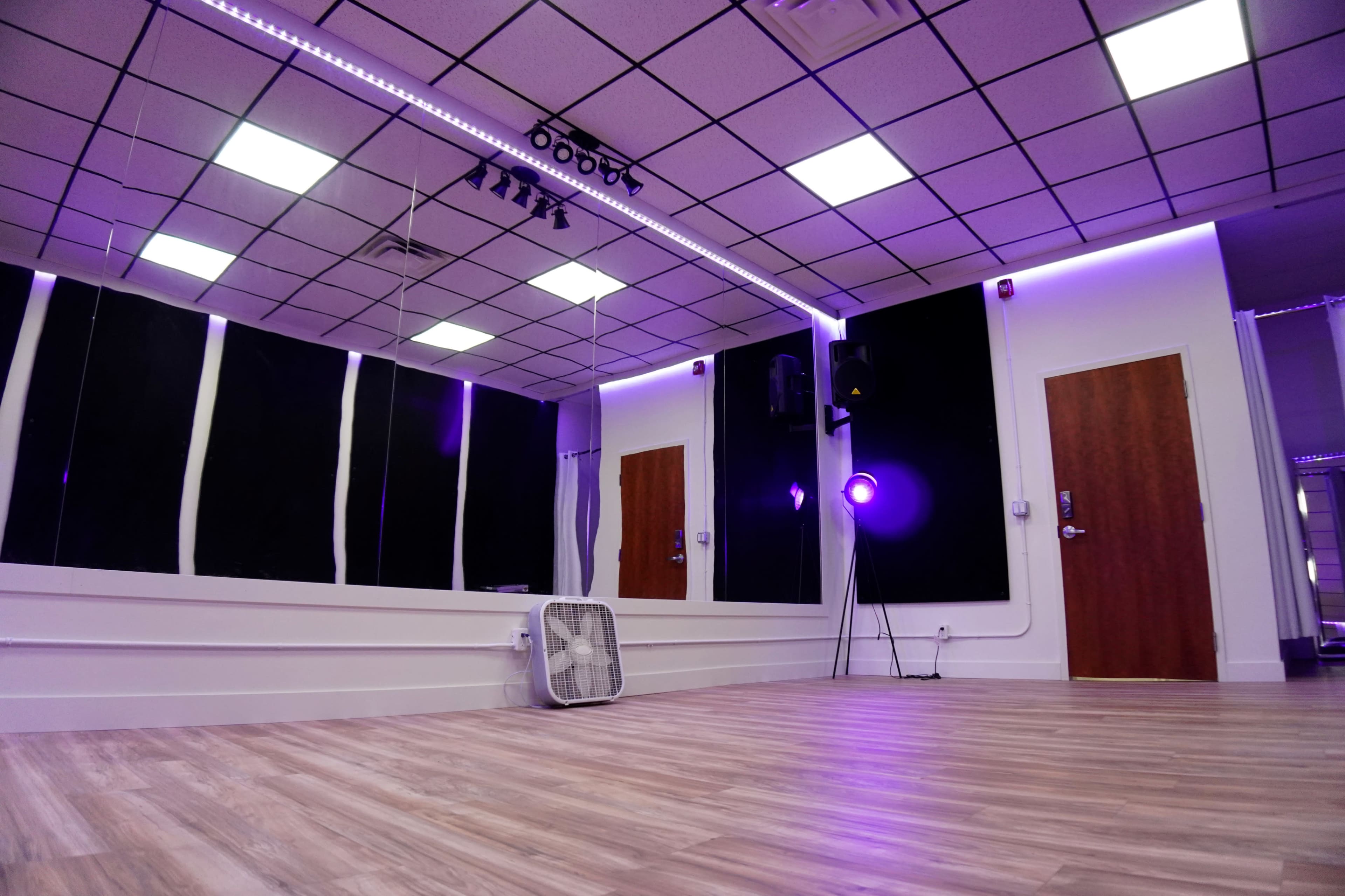 A bright, empty dance studio features a wooden floor, mirrored walls, and purple lighting.