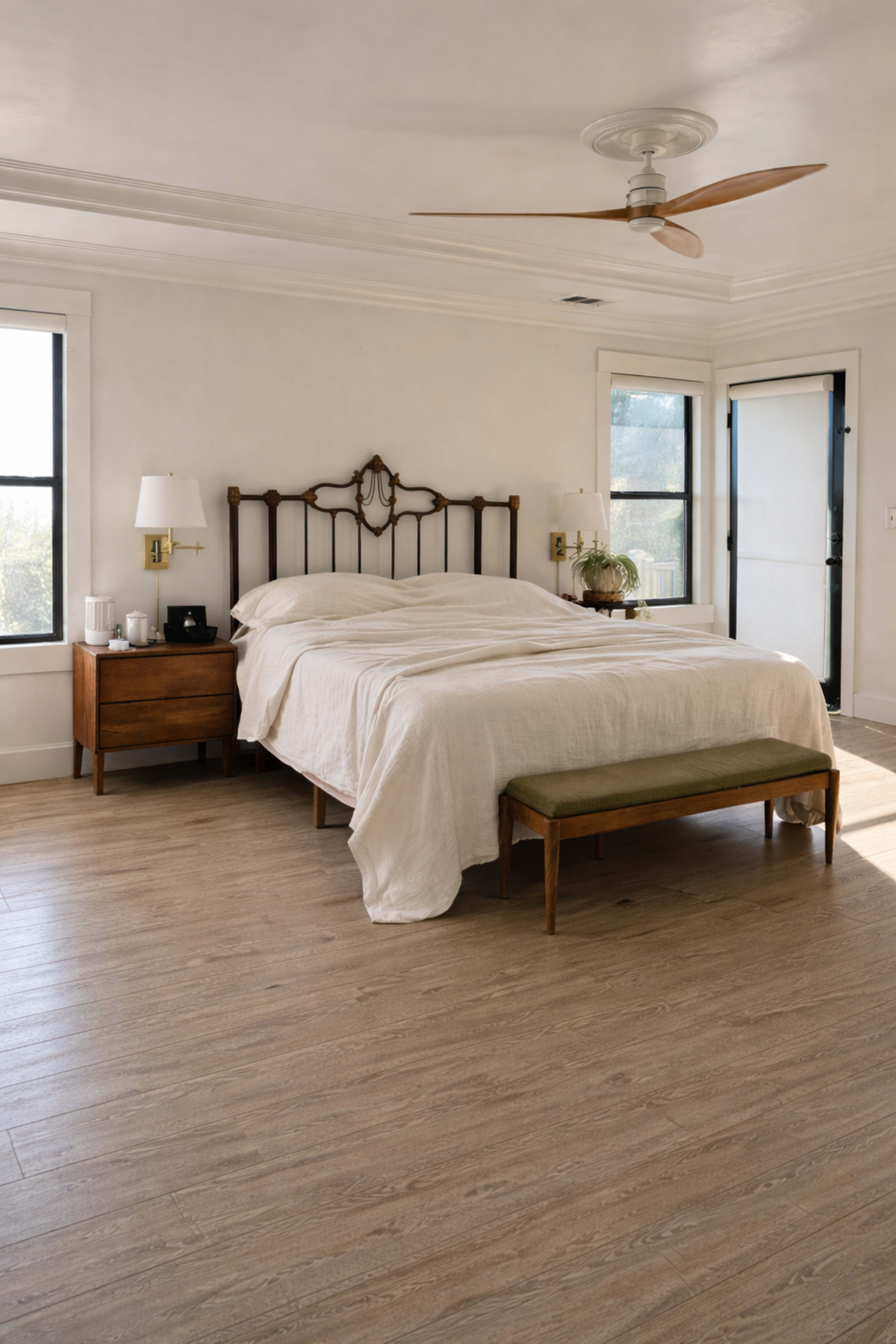 The image shows a bright bedroom featuring a metal bed frame with a light-colored duvet, a wooden nightstand with a lamp, and a bench at the foot of the bed.