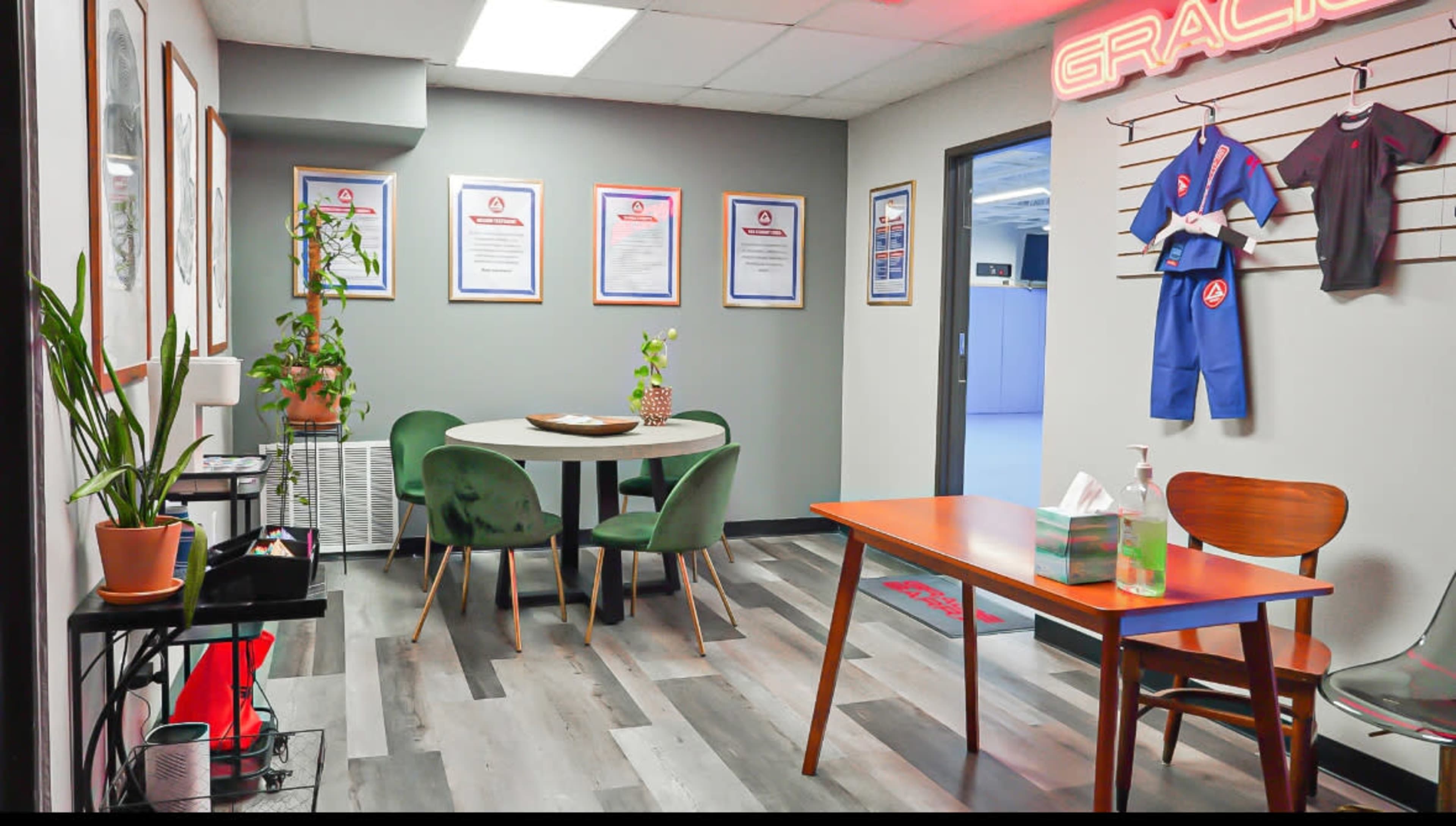 The image depicts an indoor waiting area with a round table and four green chairs, surrounded by framed certificates on the walls and a small plant in the corner.