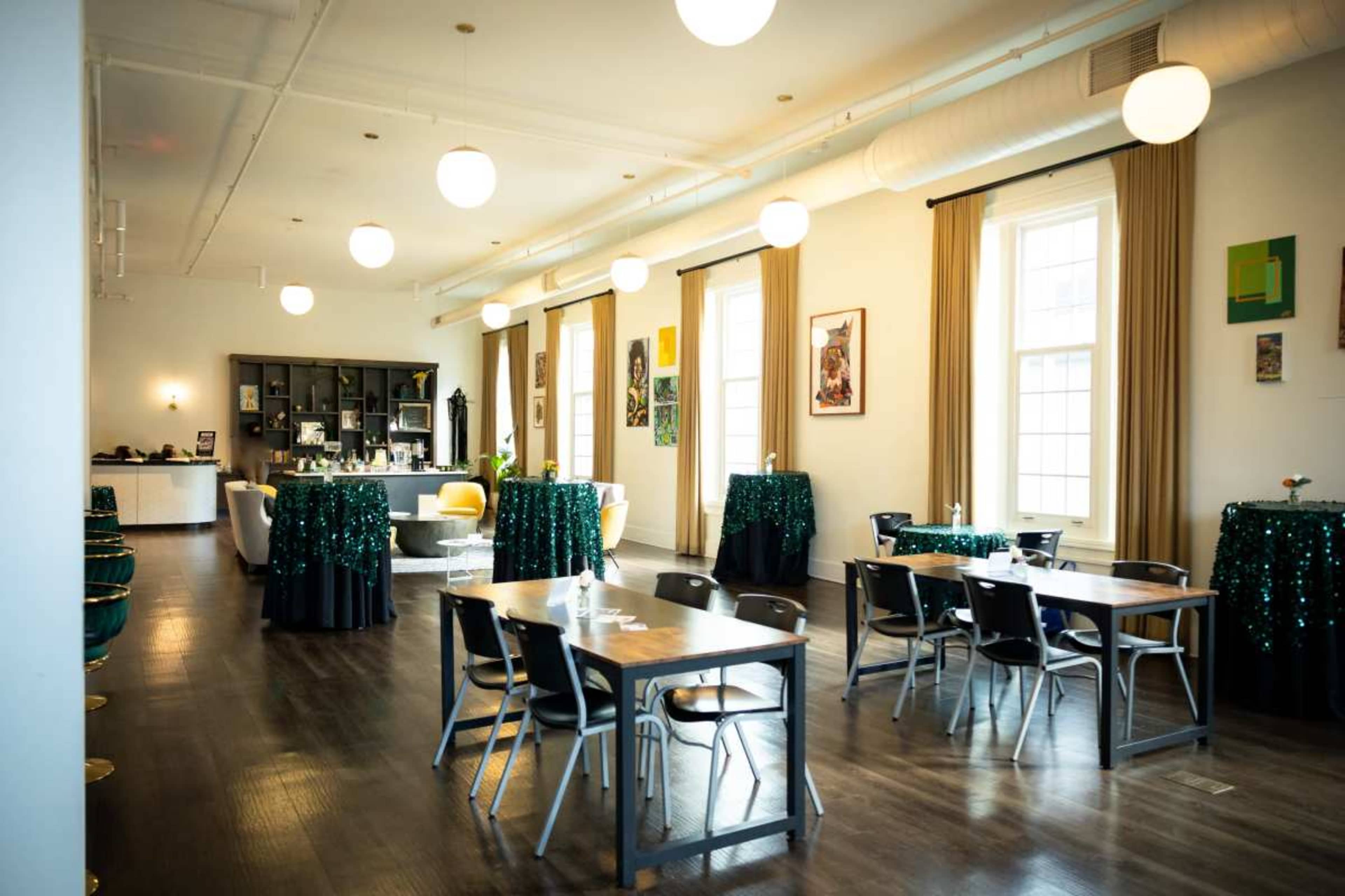 The image shows a bright, spacious event space with tables and chairs arranged, green sequin tablecloths, and large windows along the walls.