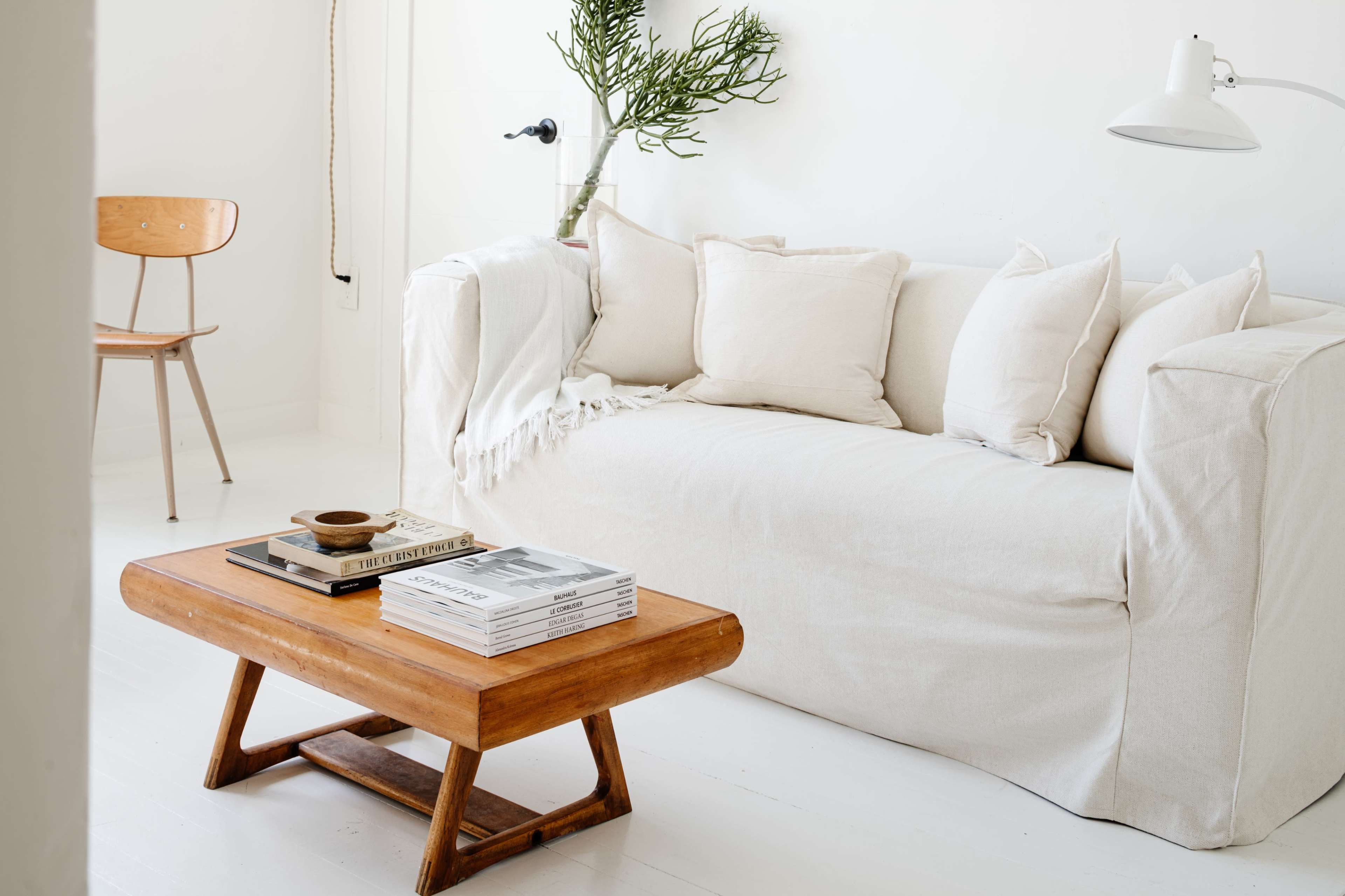 The image features a light-colored sofa with several cushions, accompanied by a wooden coffee table stacked with books, set in a minimalist living room.