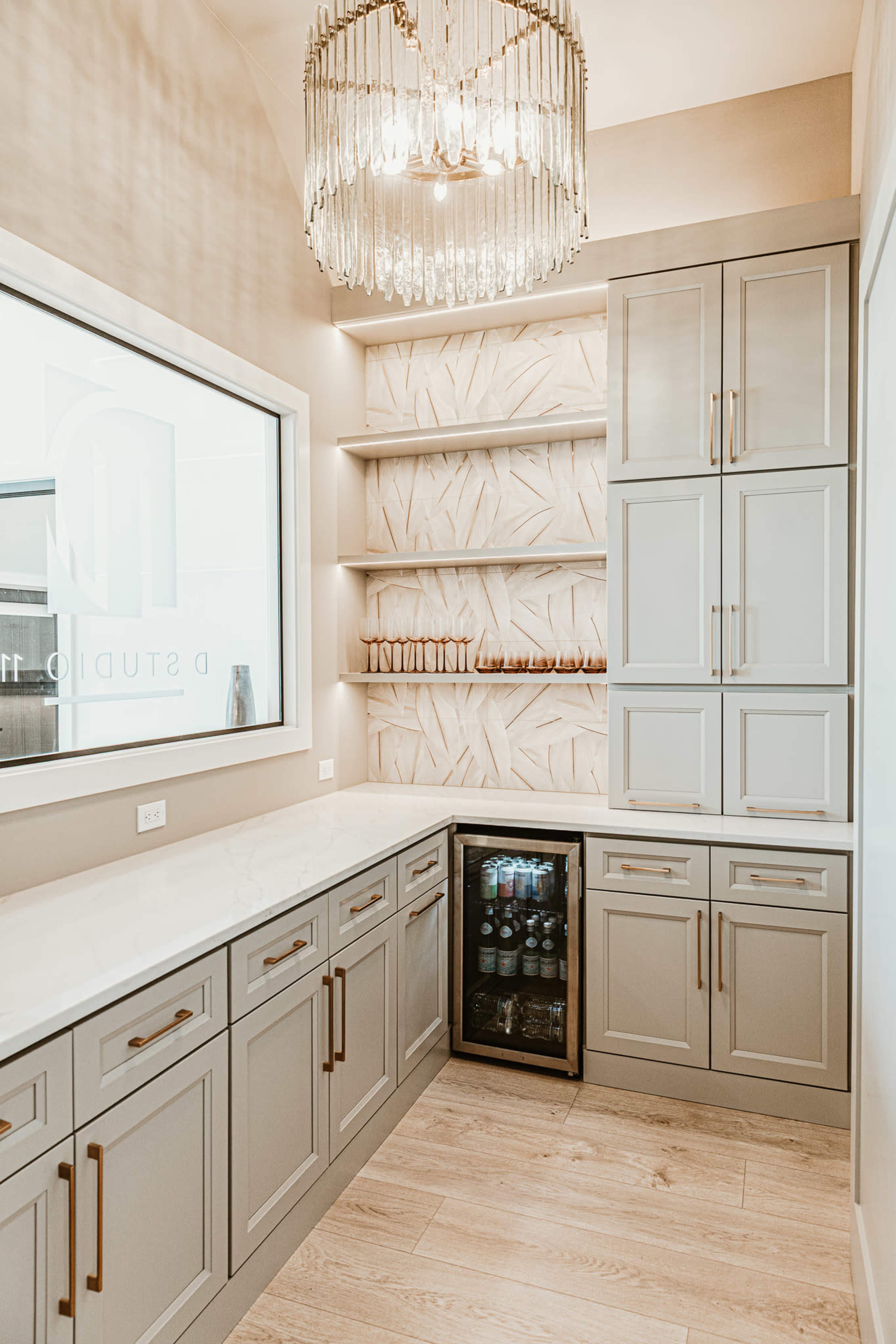 The image shows a modern kitchen corner with gray cabinetry, a small glass-front refrigerator, and a chandelier overhead, featuring a textured wall design and open shelving.