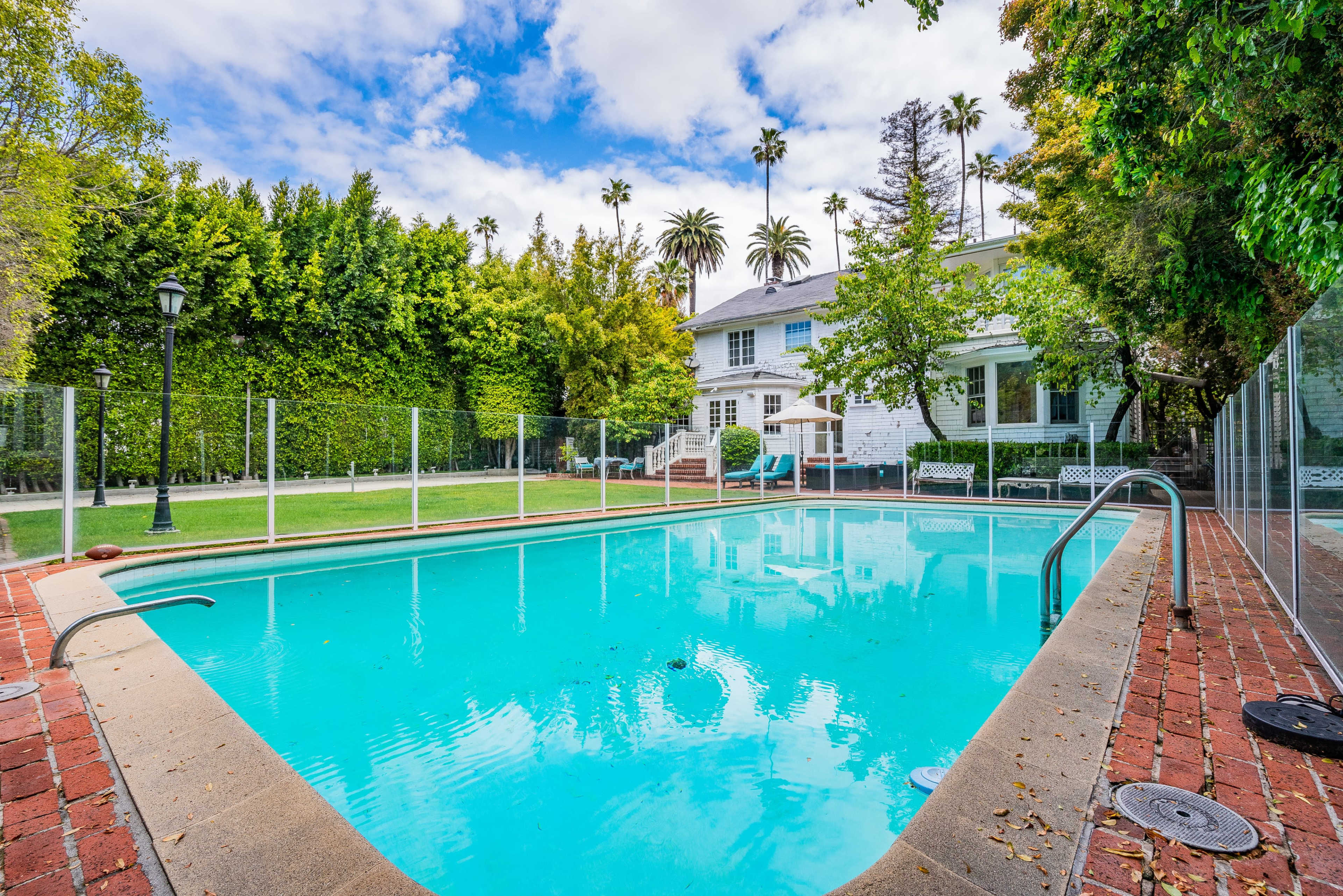 A swimming pool surrounded by a brick pathway, with a well-maintained yard and a large white house in the background.