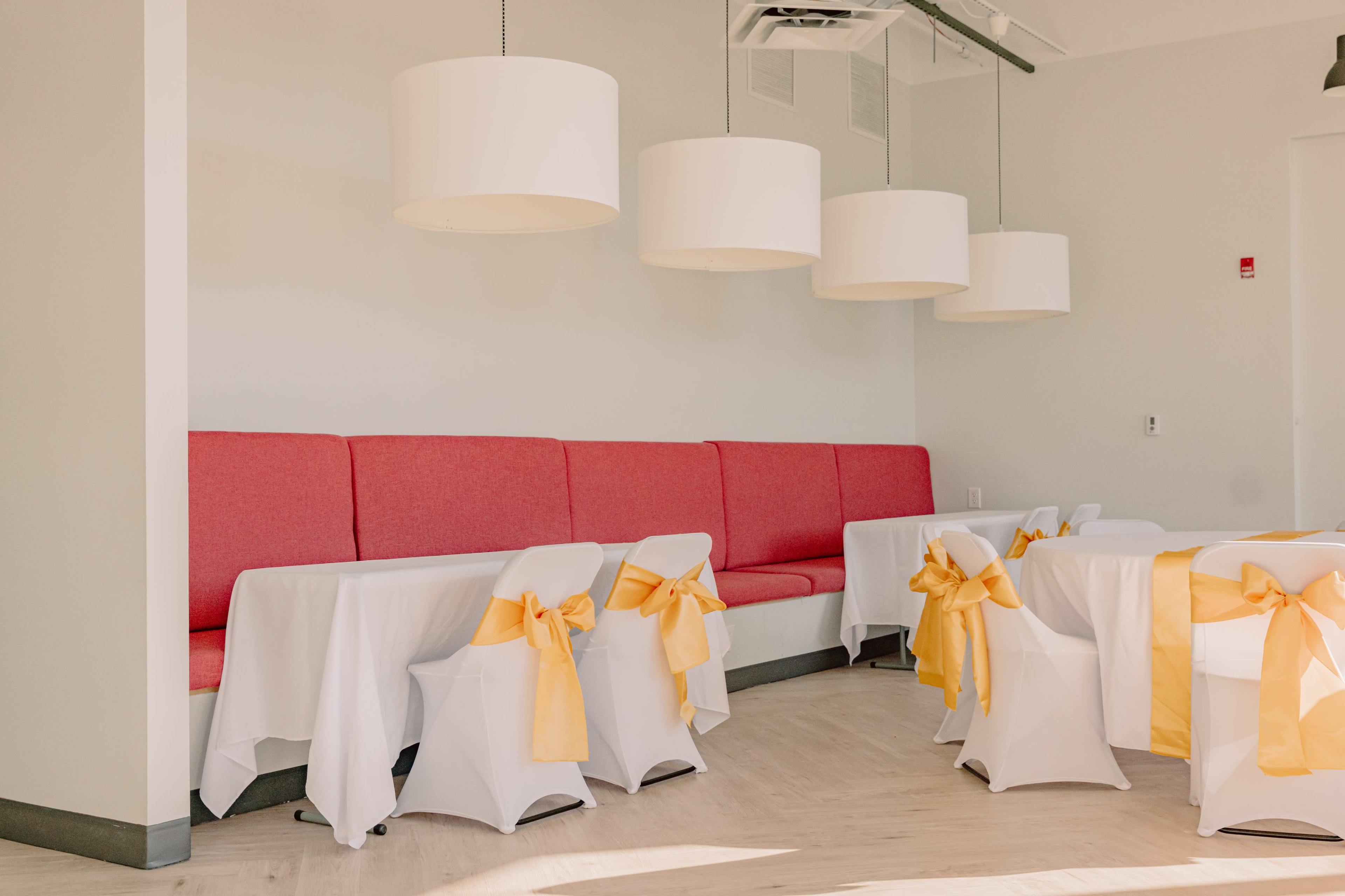 A dining area with a red upholstered bench, white tables covered in tablecloths, and yellow bows tied around the chairs.