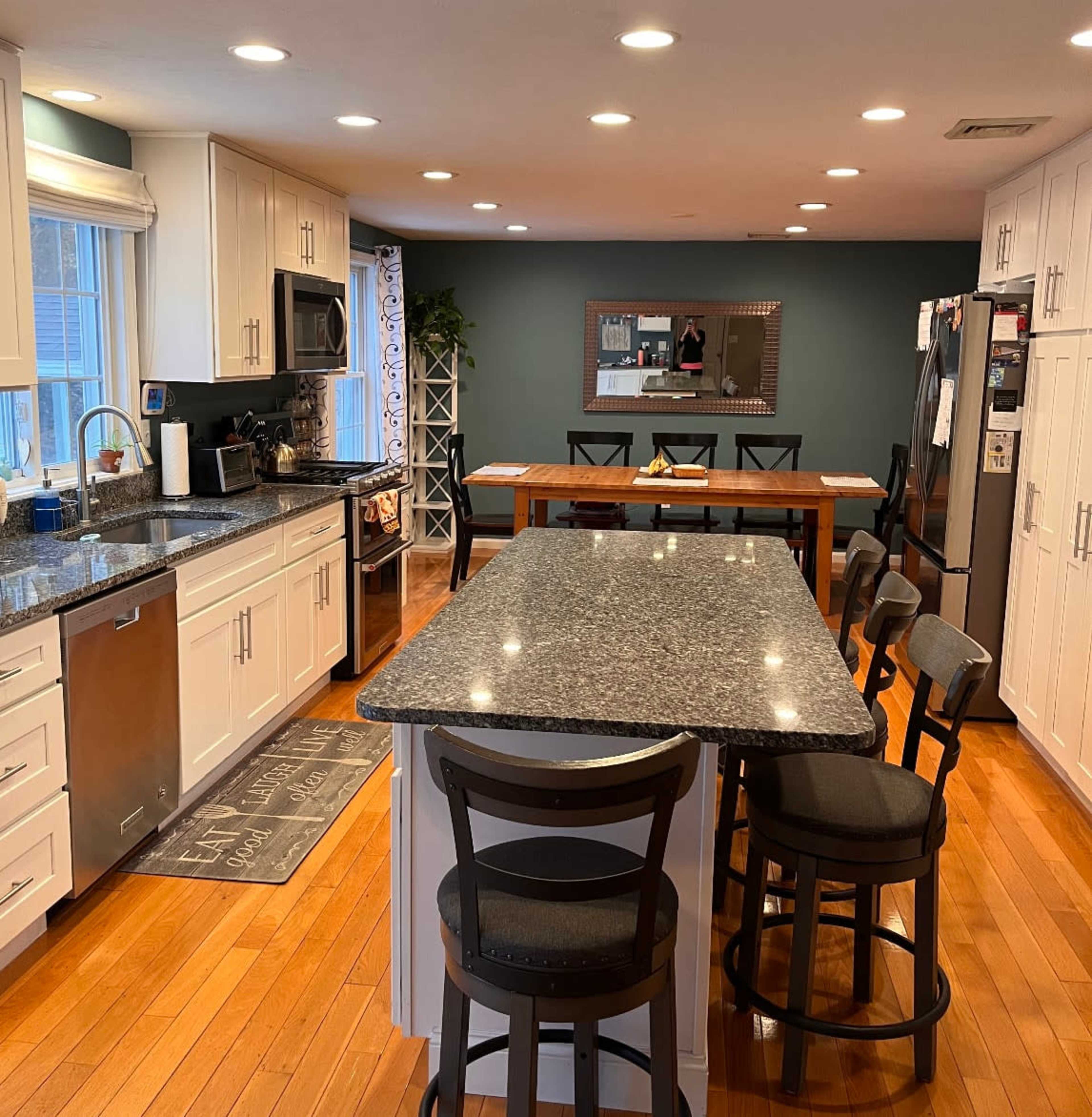 The image shows a modern kitchen featuring a large central island with bar seating, granite countertops, white cabinetry, stainless steel appliances, and a dining table in the background.