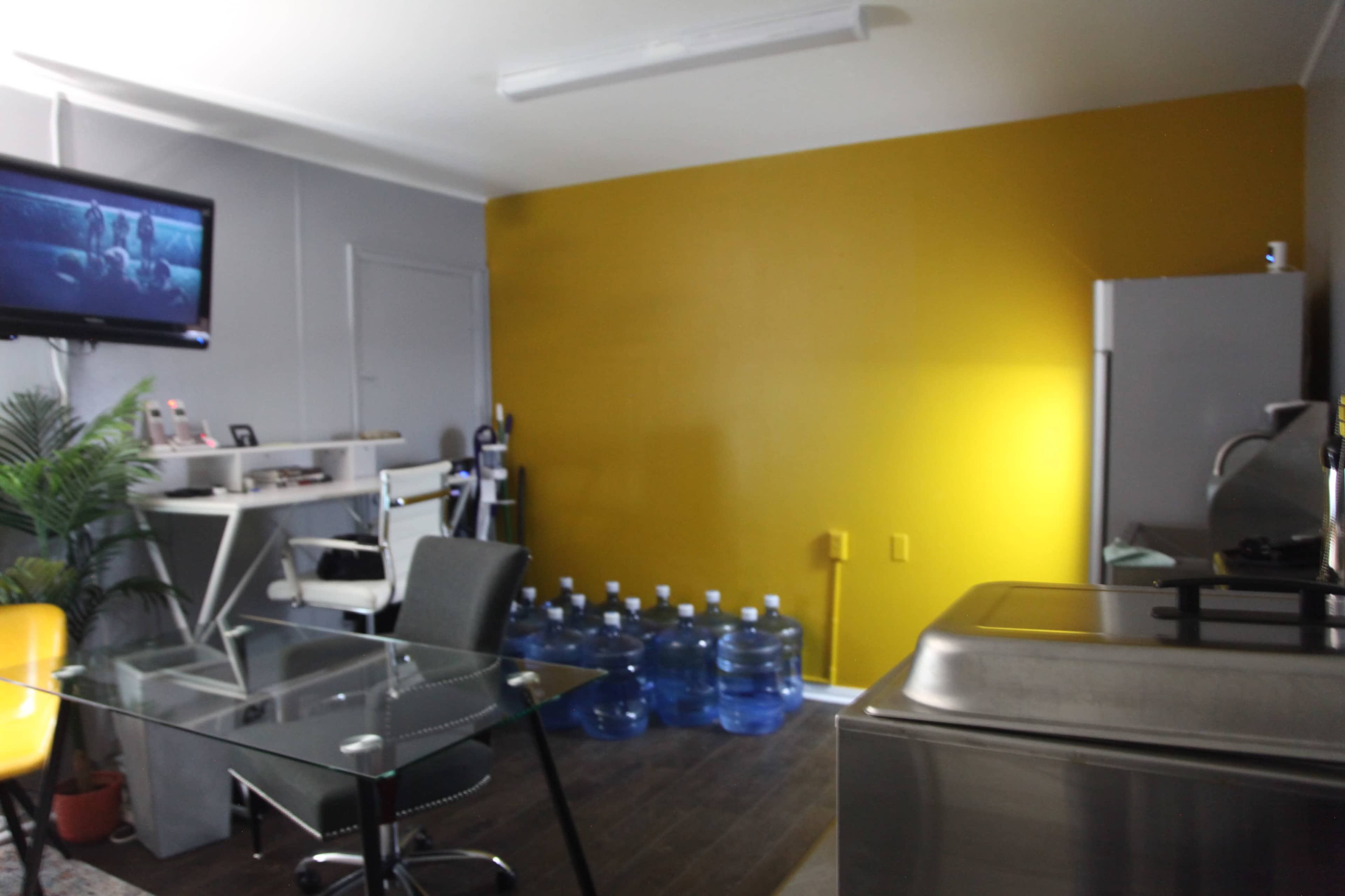 A small room features a glass table, a television mounted on the wall, a yellow accent wall, and several blue water bottles lined up against the side.