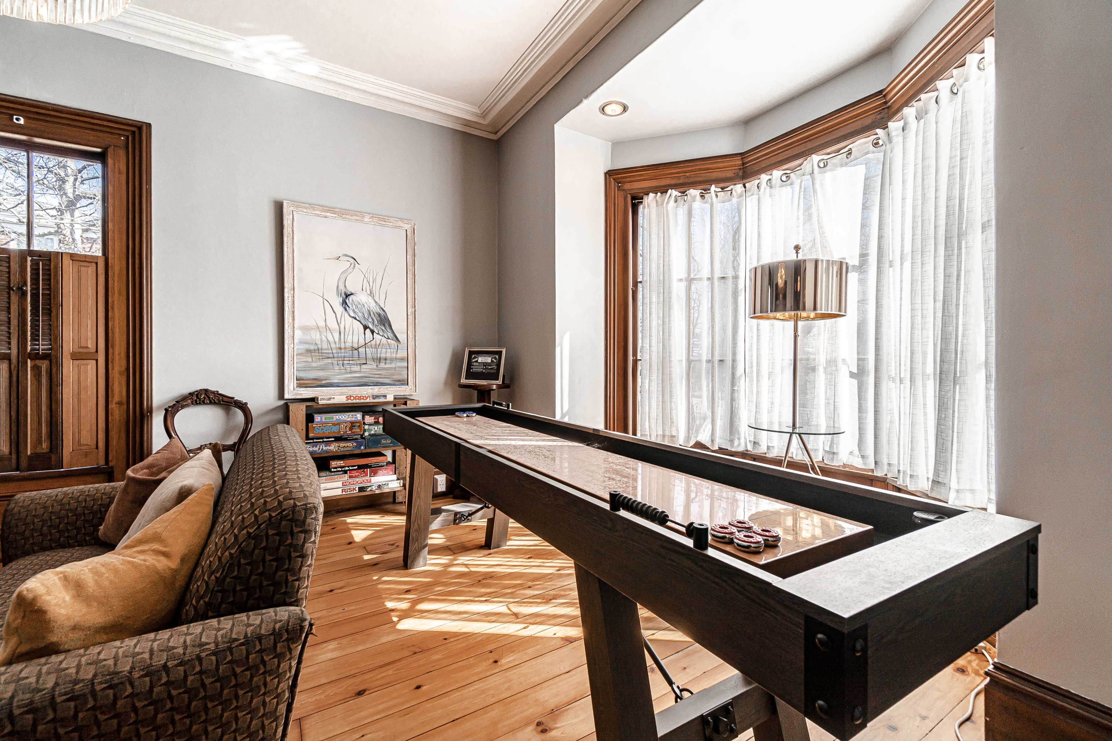 A sunlit room features a shuffleboard table next to a comfortable chair and large windows with sheer curtains.