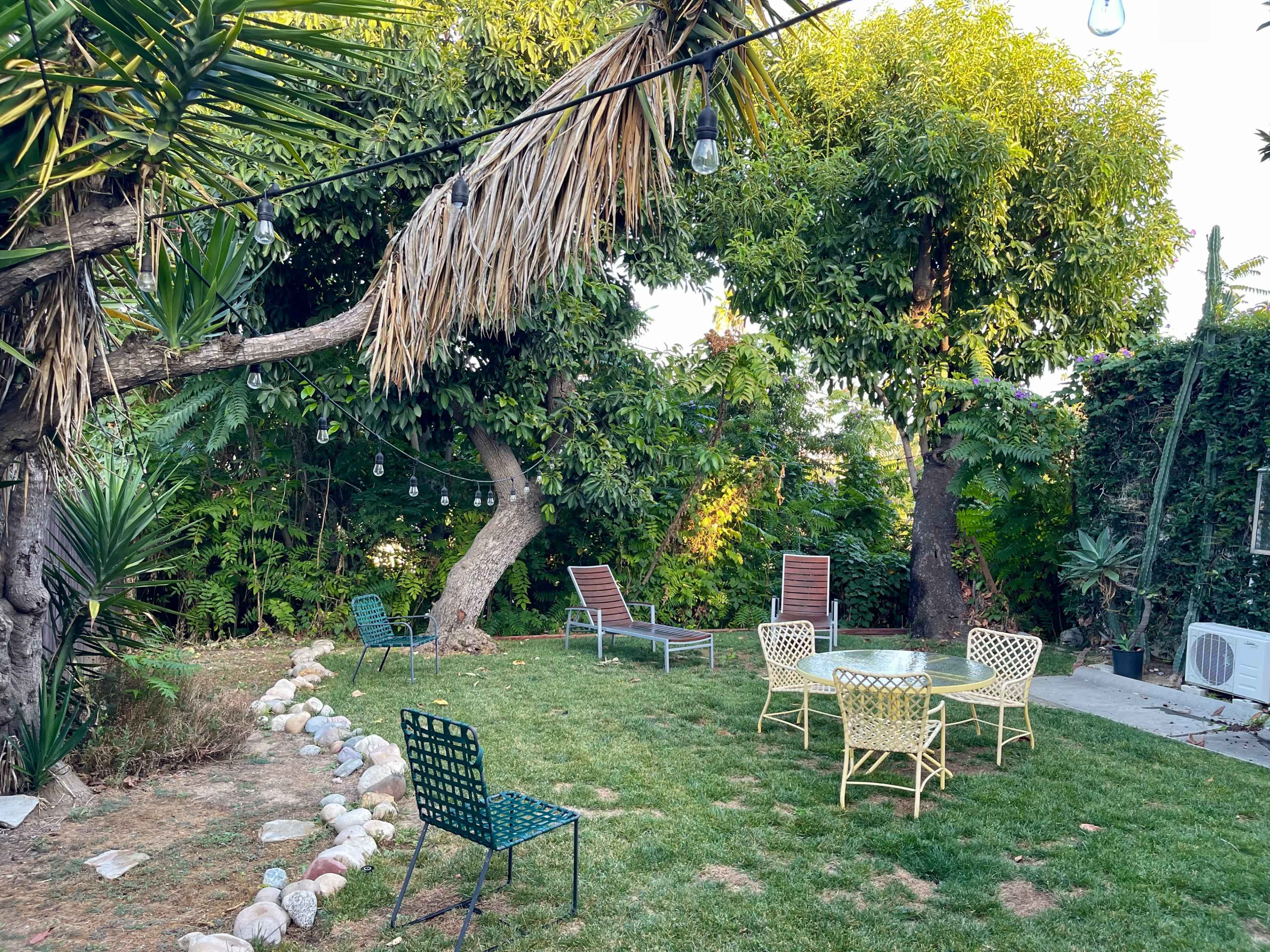 A backyard garden with green grass, lounge chairs, and a table surrounded by trees and decorative lights.