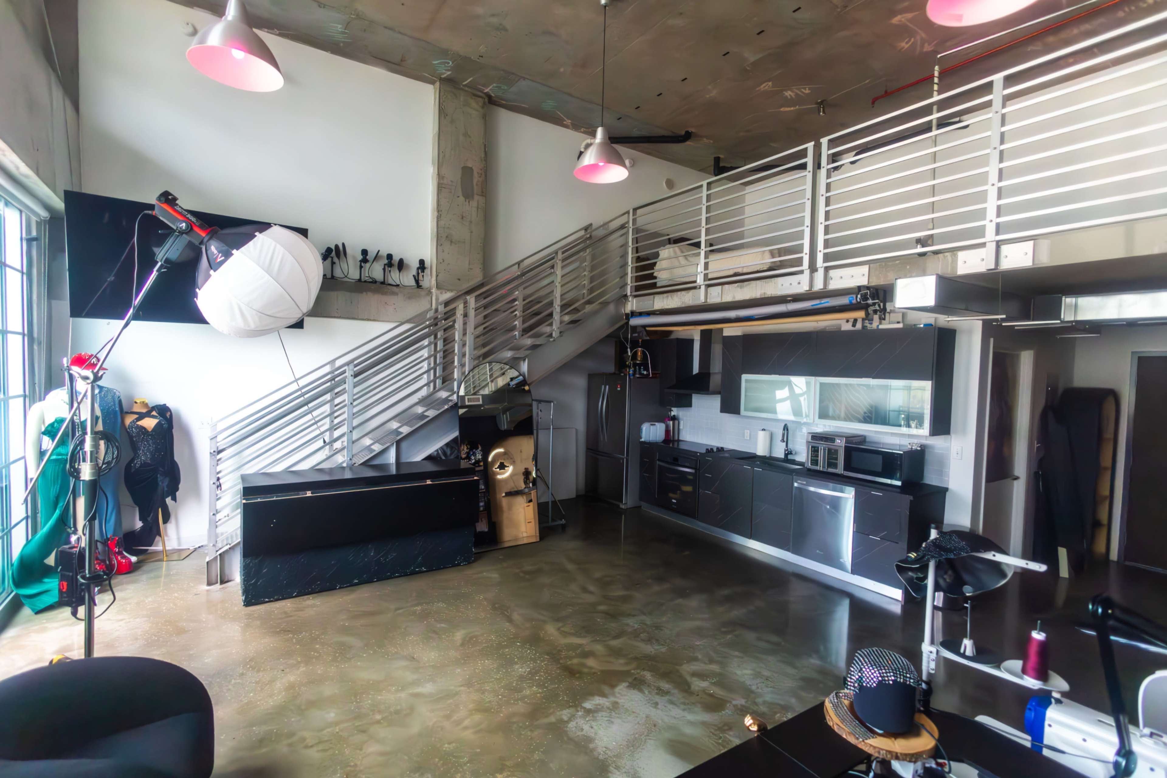 The image shows a modern industrial-style loft with an open floor plan, featuring a kitchen area, a staircase with metal railings, and various equipment and furnishings scattered throughout.
