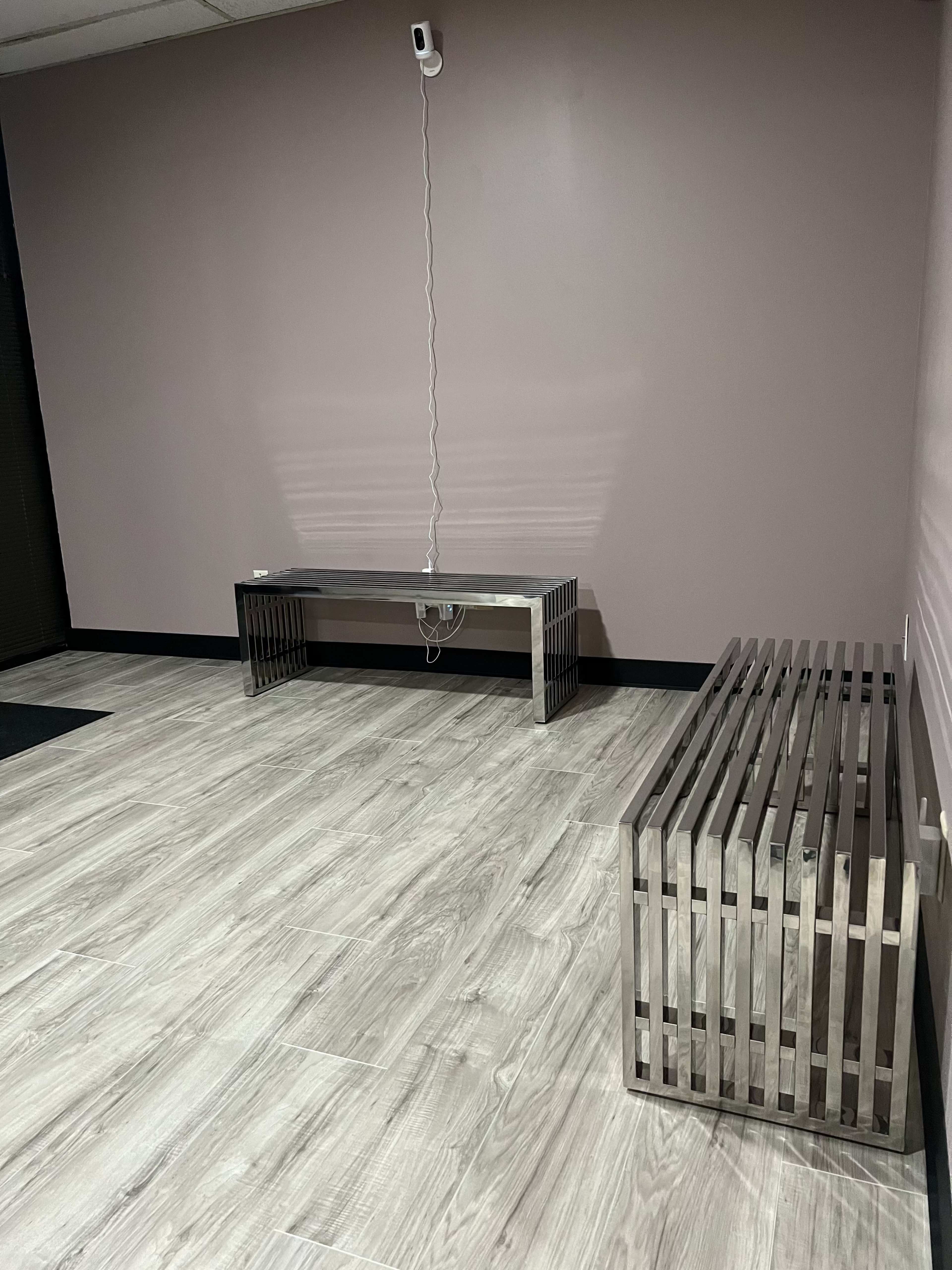 A simple interior space featuring two modern metallic benches on a wooden floor with a plain wall in the background.