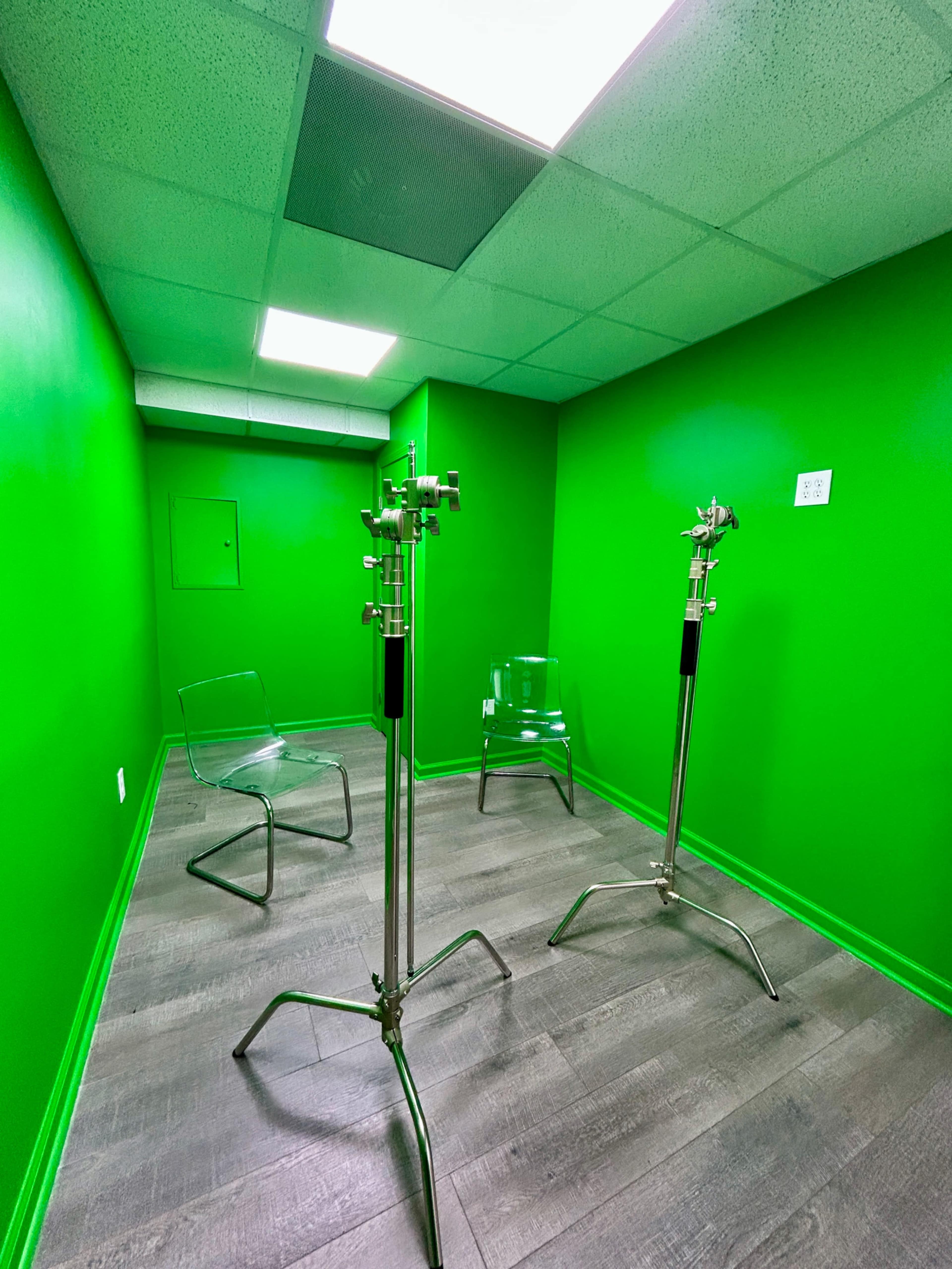 The image shows a small room with bright green walls, two metal light stands, and two transparent chairs on a wooden floor.