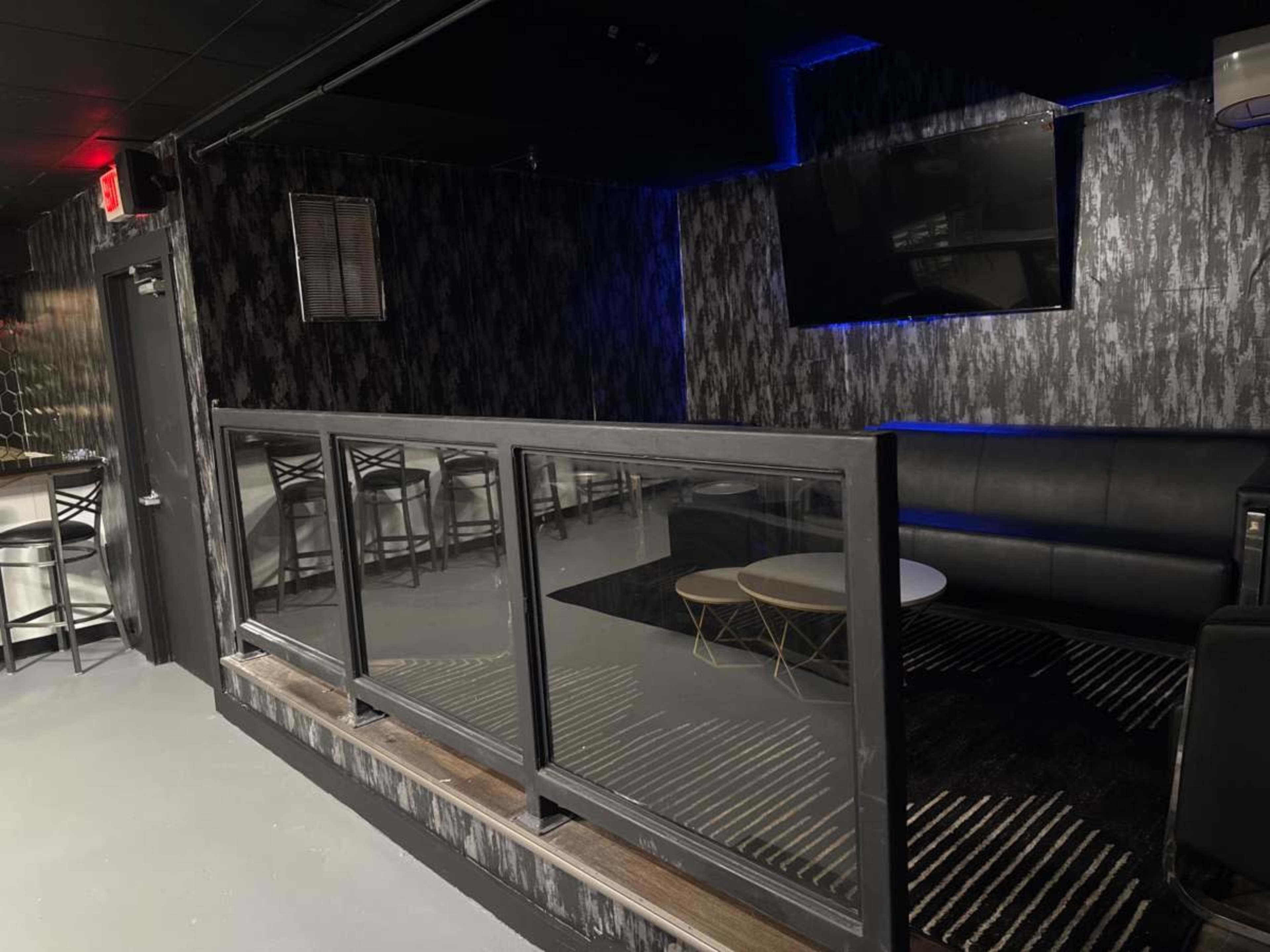 The image shows a dimly lit lounge area featuring a sectional sofa, a coffee table, and a small railing partition, with a television mounted on the wall.