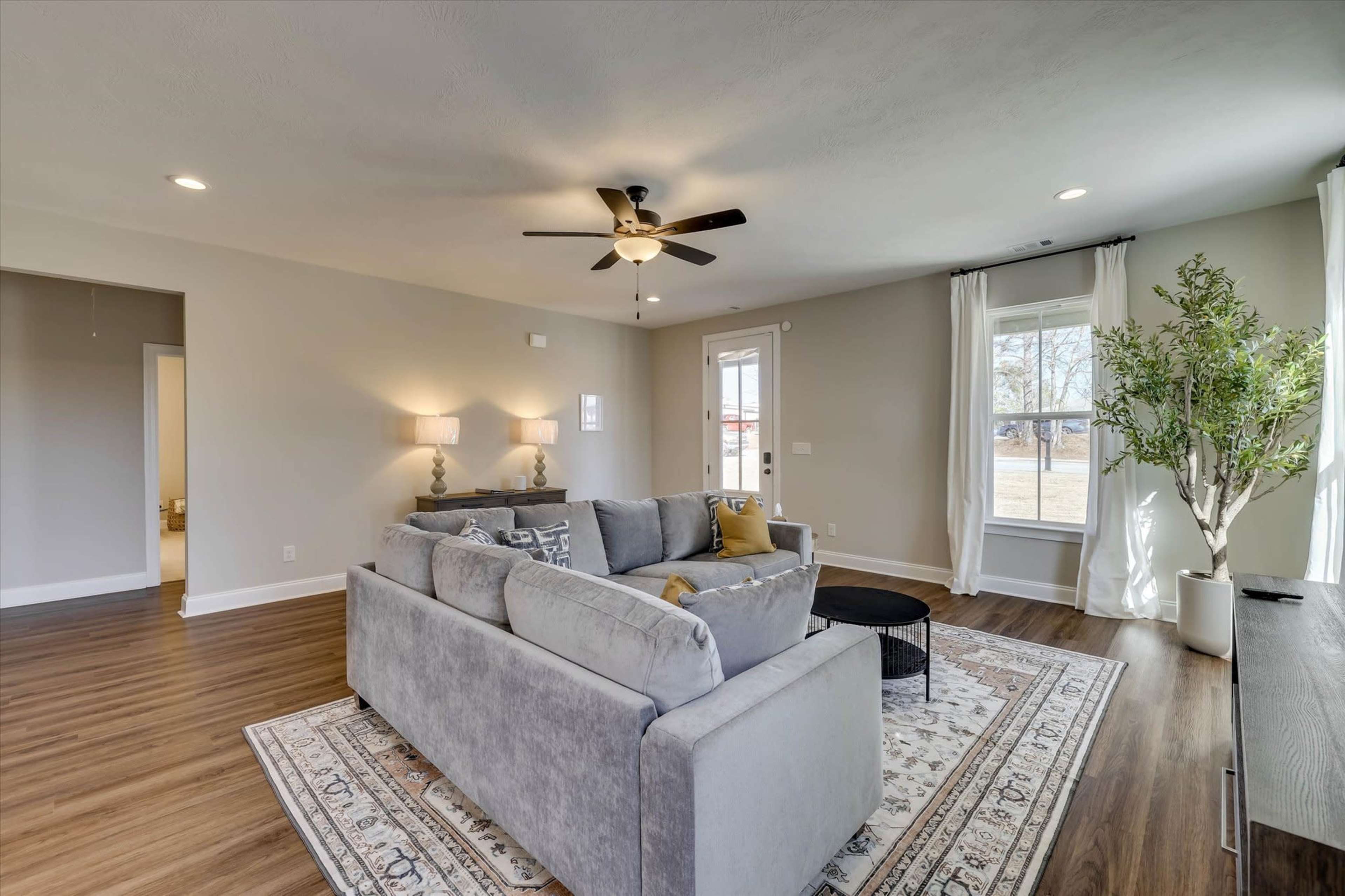 The image shows a spacious living room featuring a gray sectional sofa, a round black coffee table, a ceiling fan, and large windows with sheer curtains, complemented by a decorative plant in one corner.