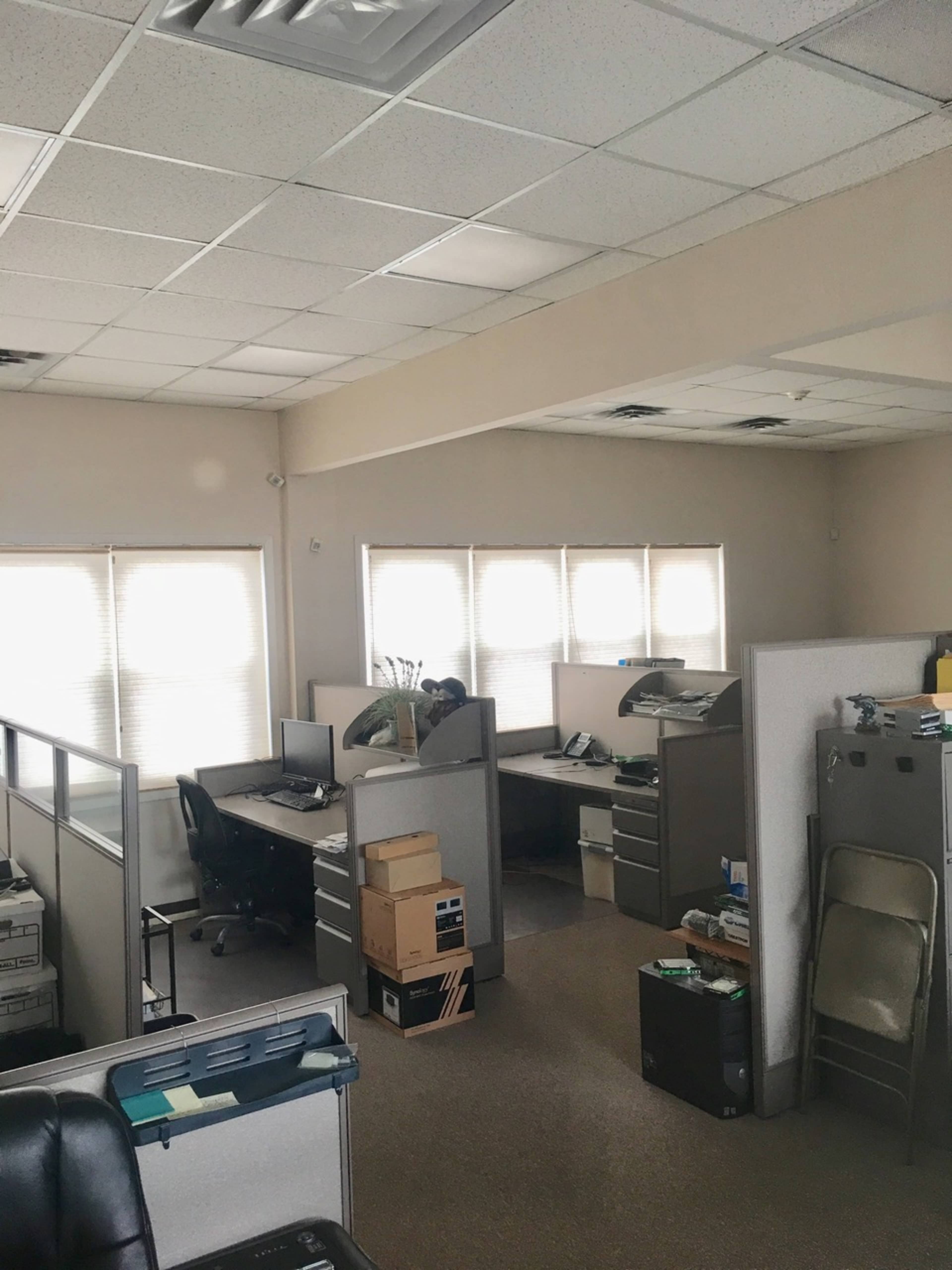 A spacious office area with several cubicles, each containing a desk and office equipment, illuminated by natural light from adjacent windows.