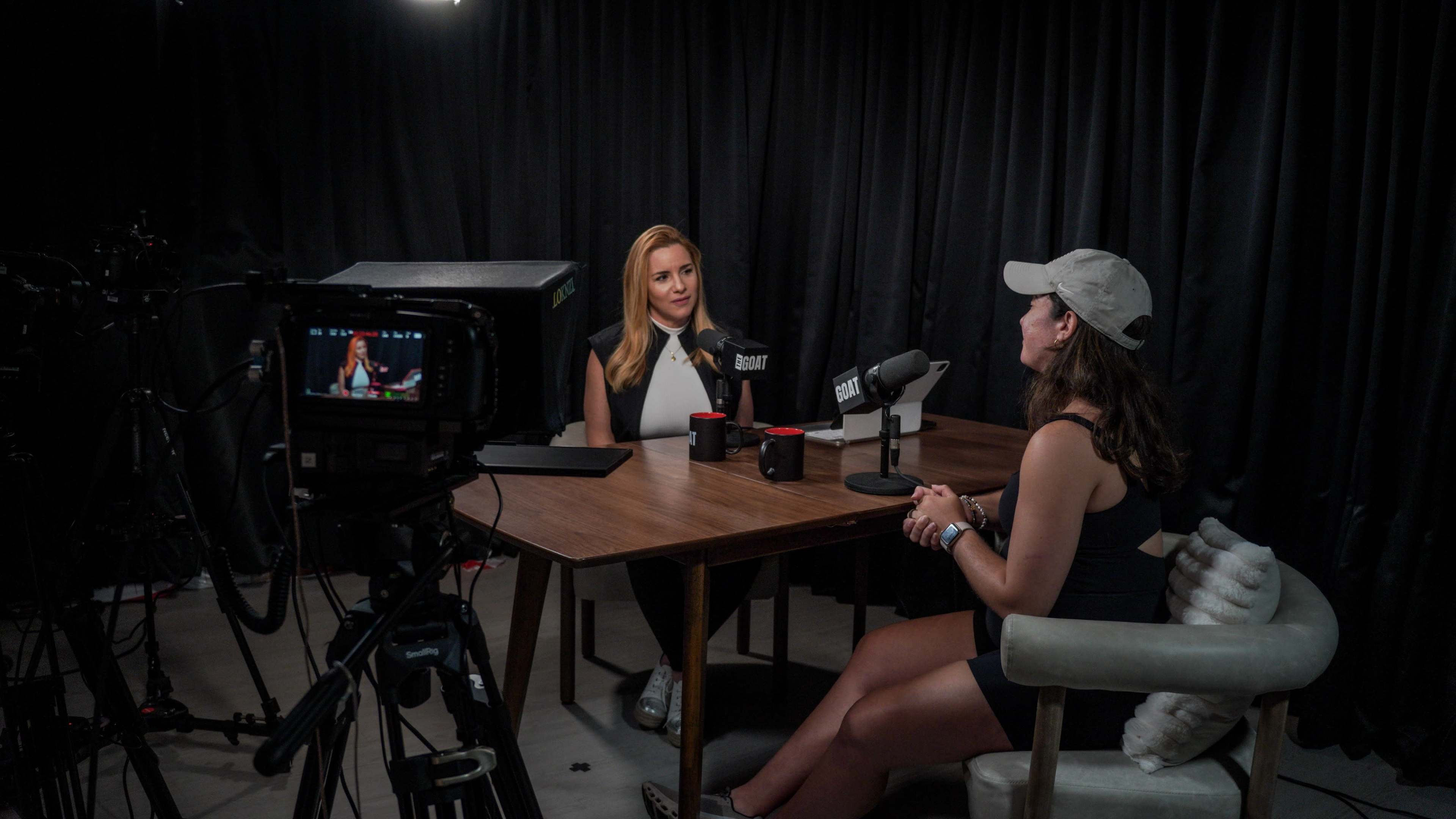 Two women sit at a table in a podcast studio, with cameras and microphones set up around them.