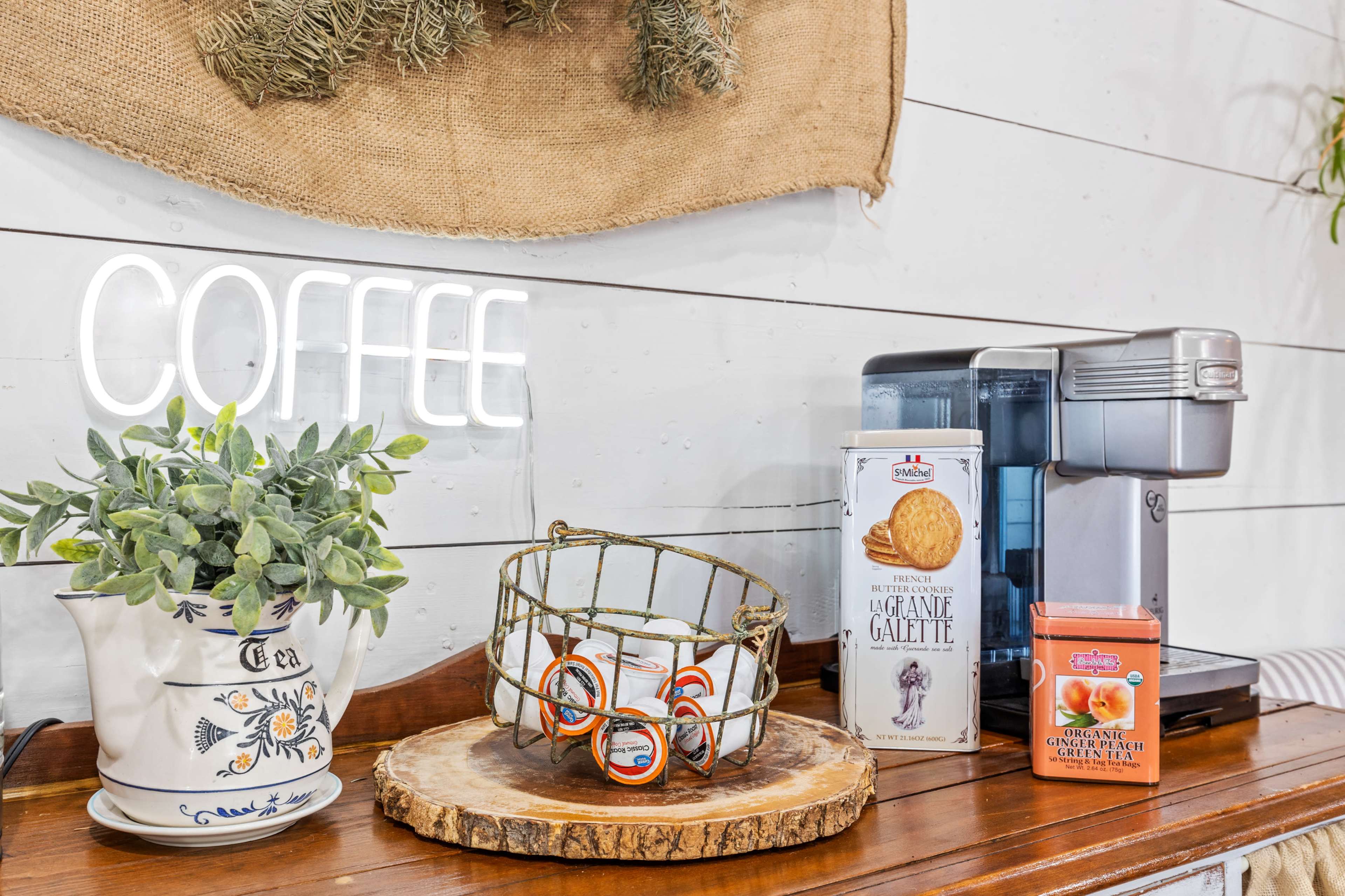 A coffee station displays a neon "COFFEE" sign, a coffee maker, assorted snacks, and a potted plant on a wooden countertop.