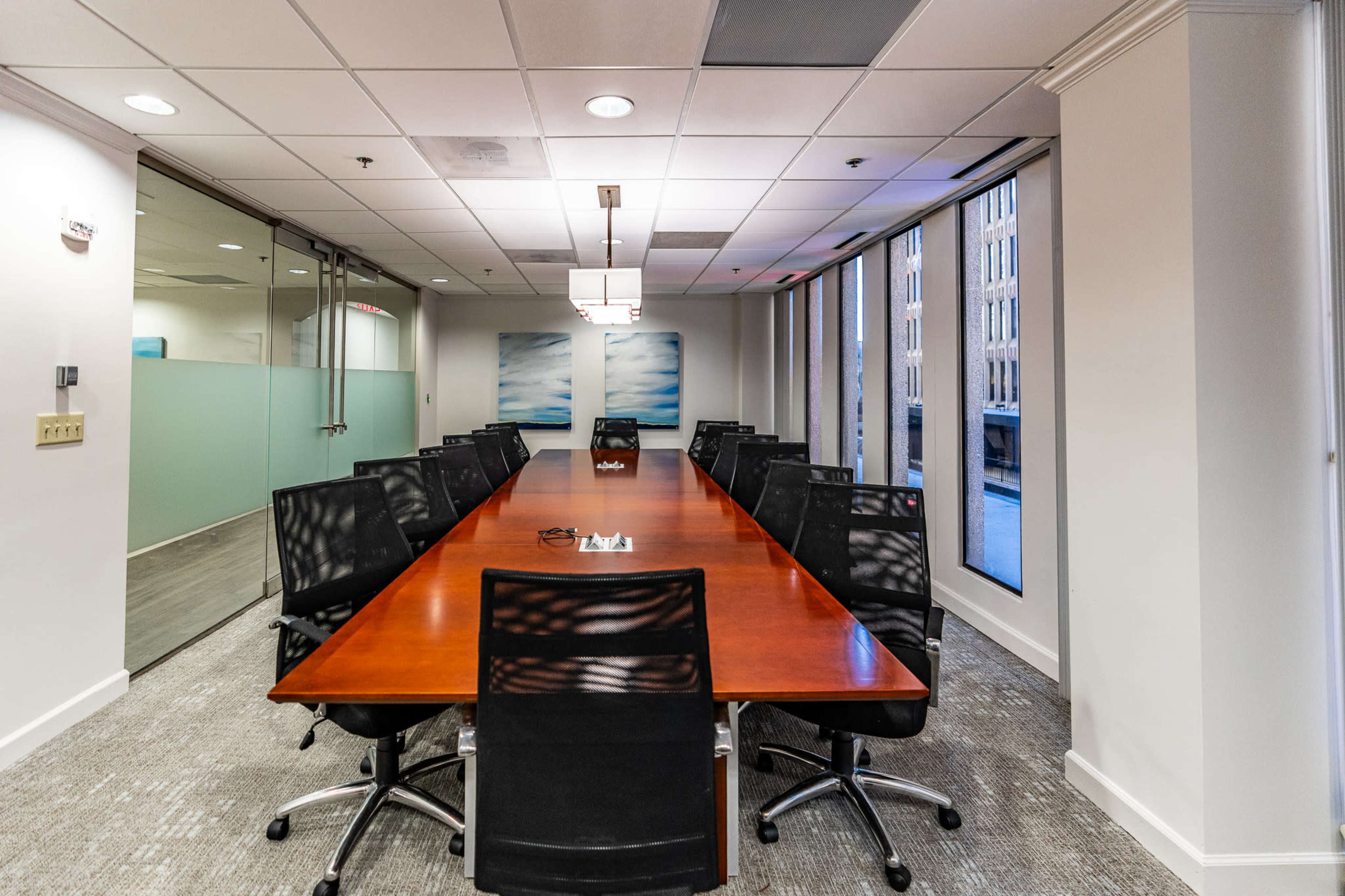 A long wooden conference table is surrounded by black chairs in a well-lit meeting room with large windows.