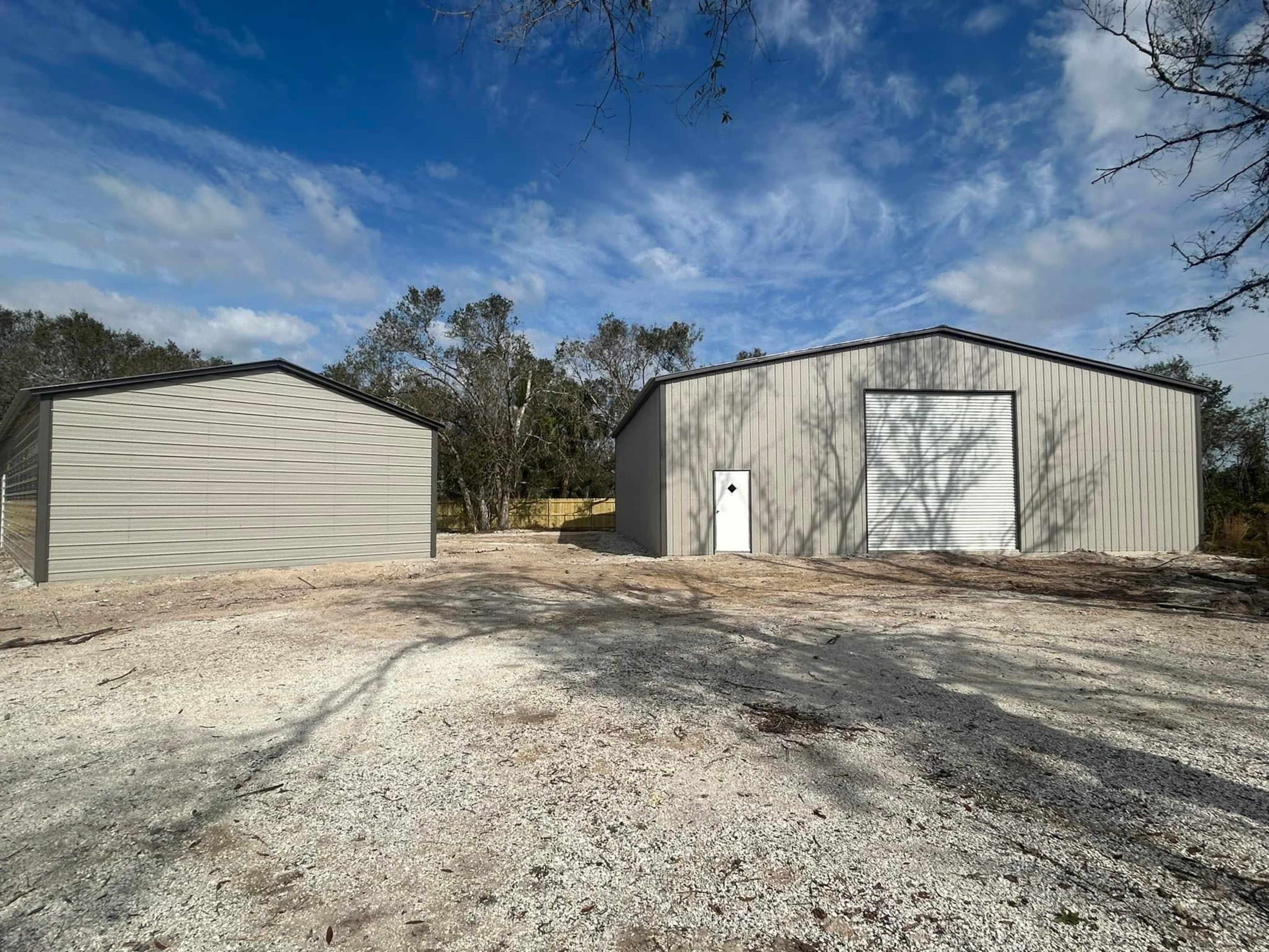 Two metal buildings on a gravel lot, surrounded by sparse trees and a partly cloudy sky.