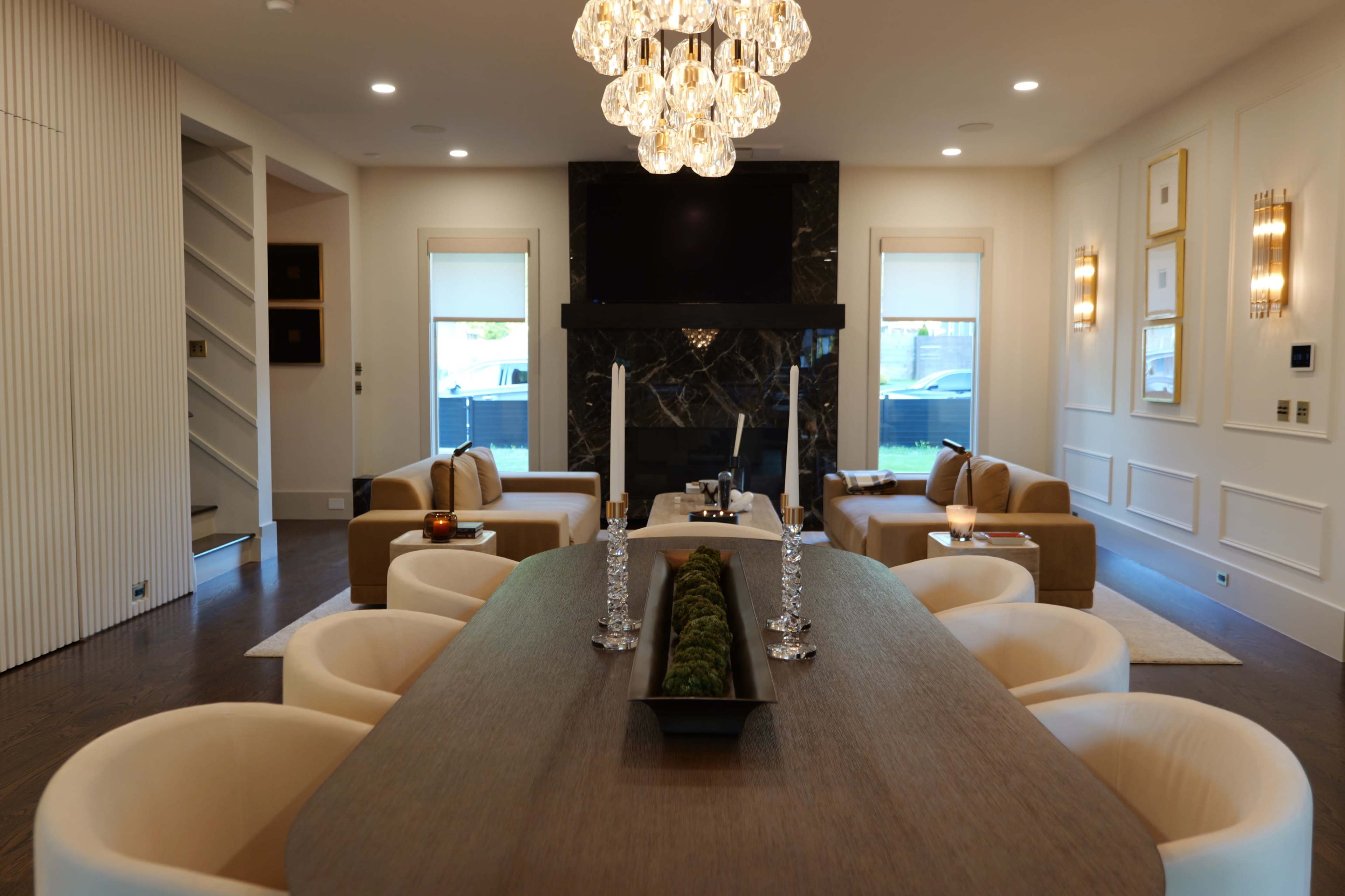 A modern living and dining area features a large wooden dining table, plush seating arrangements, a decorative centerpiece, and a statement chandelier.