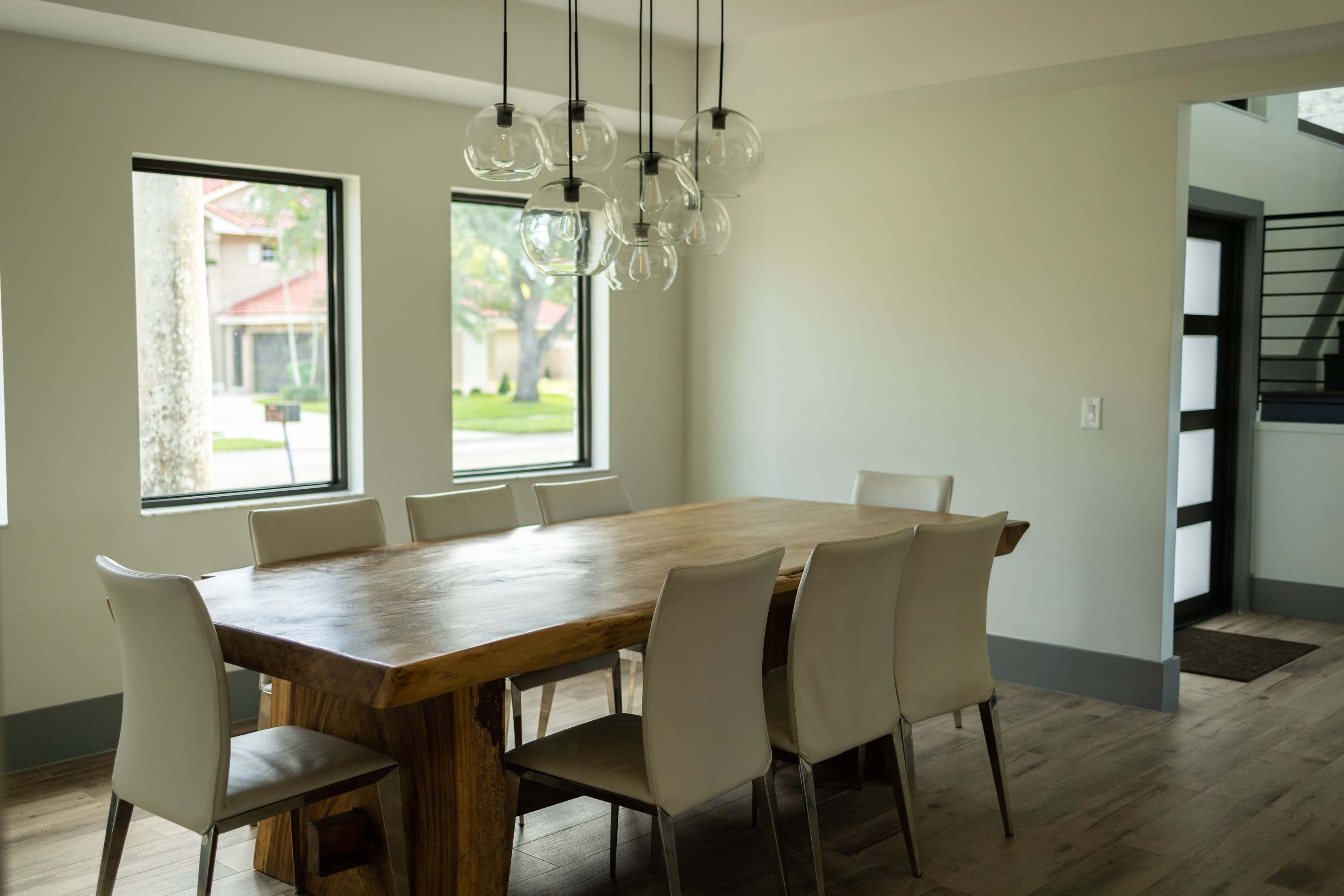 A wooden dining table with eight light-colored chairs is set in a brightly lit room featuring large windows and modern pendant lighting.