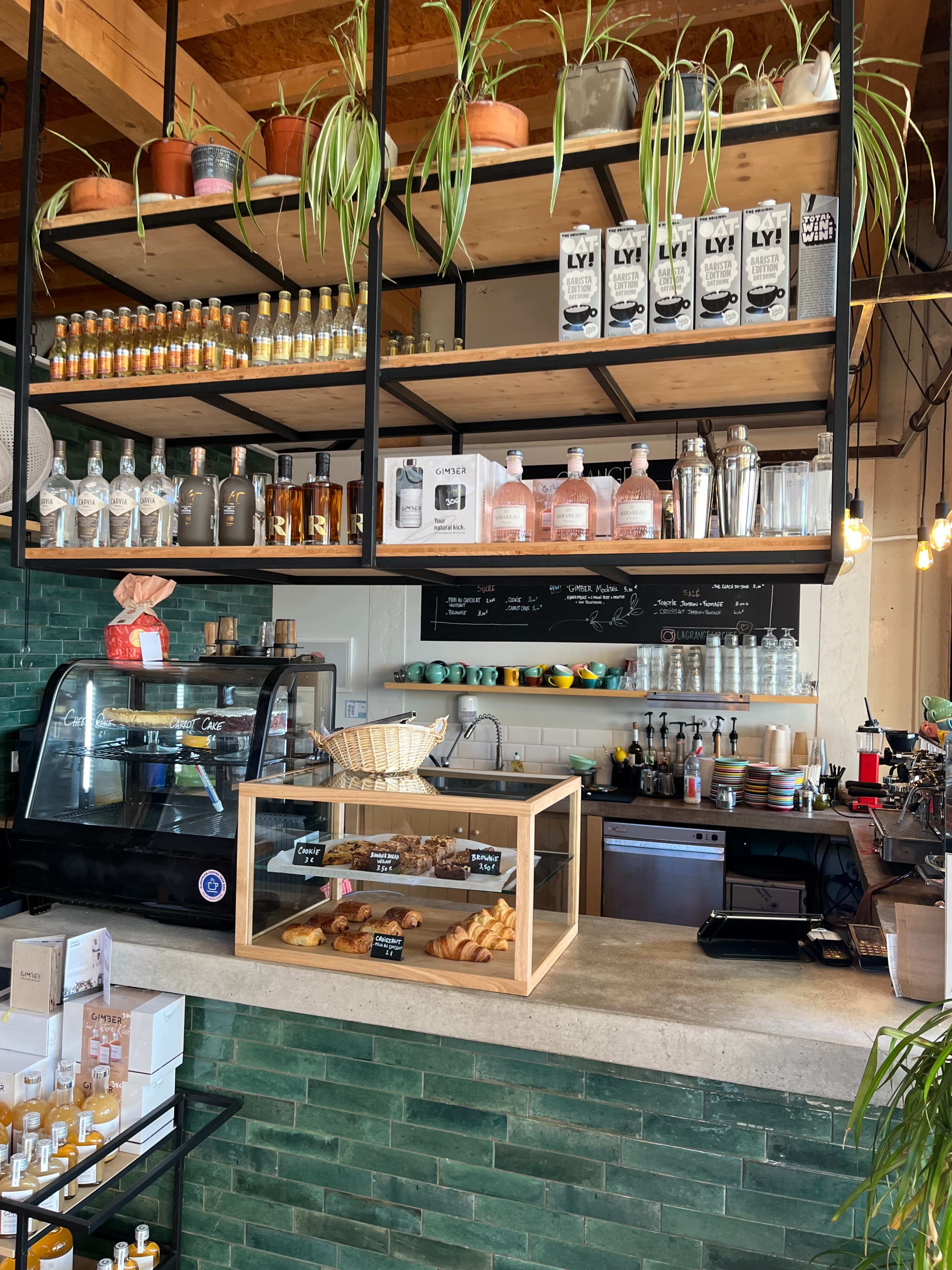 The image shows a café interior with shelves displaying beverages and snacks, a glass display case containing baked goods, and a counter equipped with coffee-making equipment.