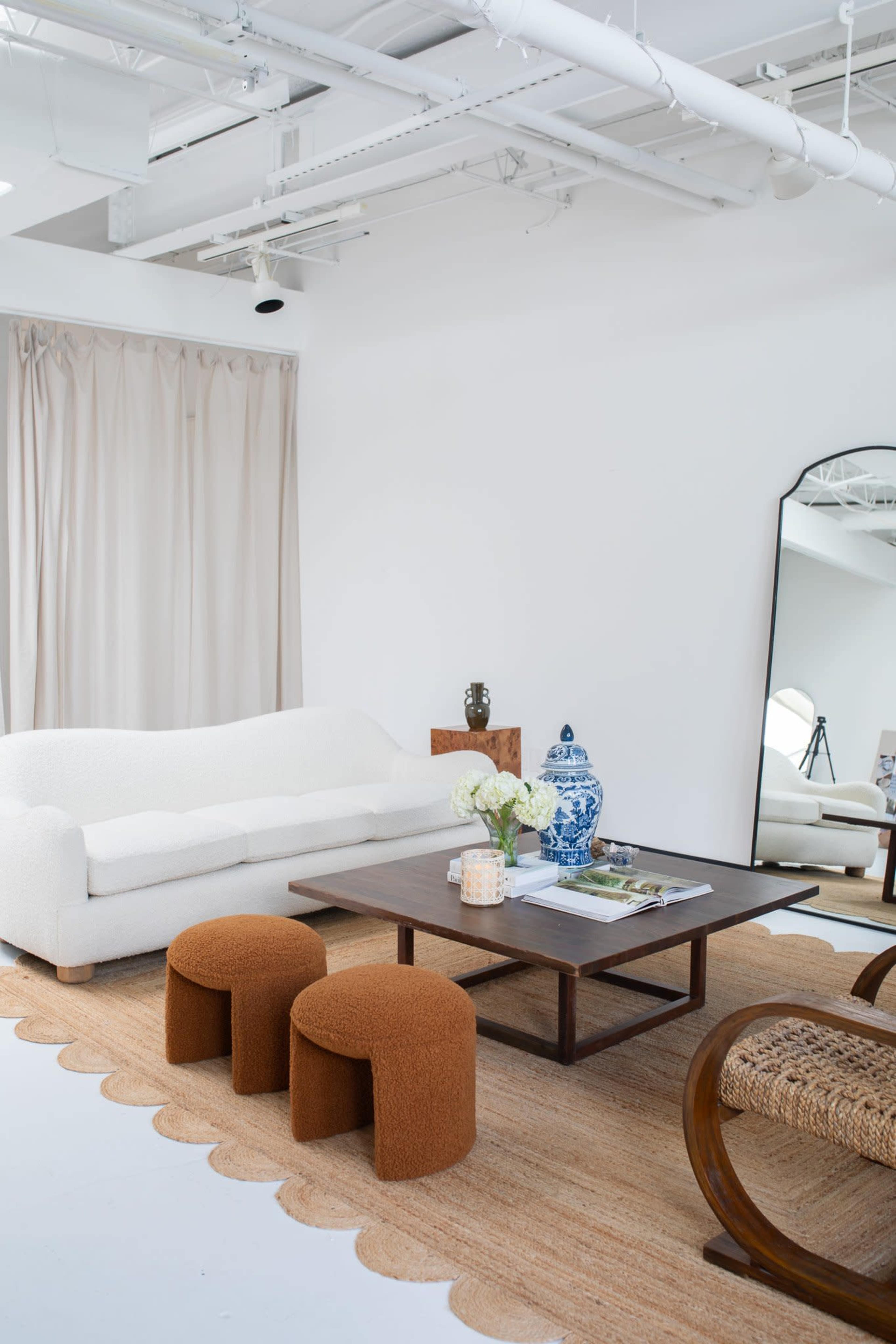 A cozy living room features a white sofa, two rounded brown stools, a wooden coffee table, and a large mirror against a bright white wall.