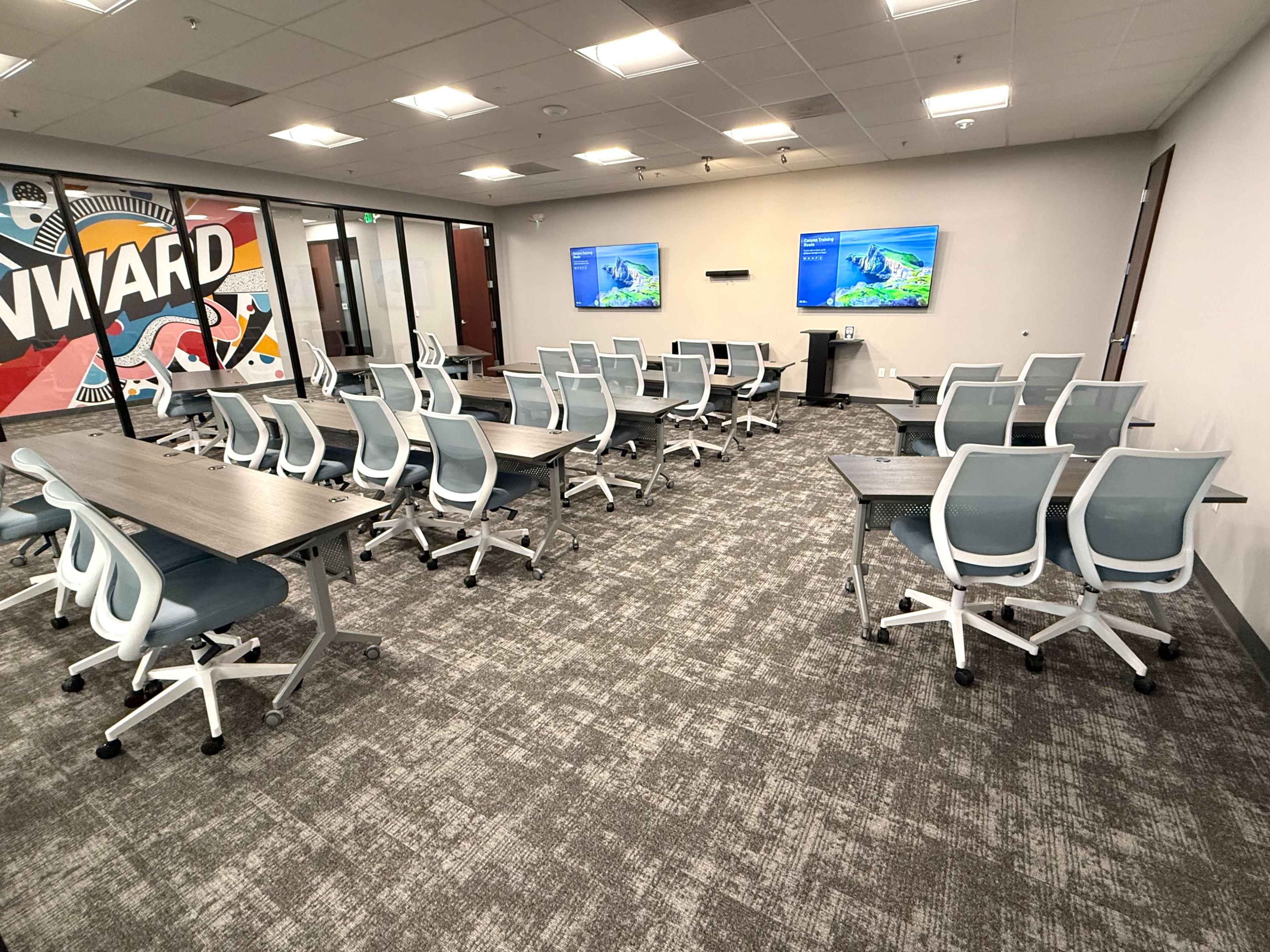 The image shows a modern, empty classroom setup with rows of movable tables and chairs, two wall-mounted screens displaying blue graphics, and a colorful mural featuring the word "ONWARD."