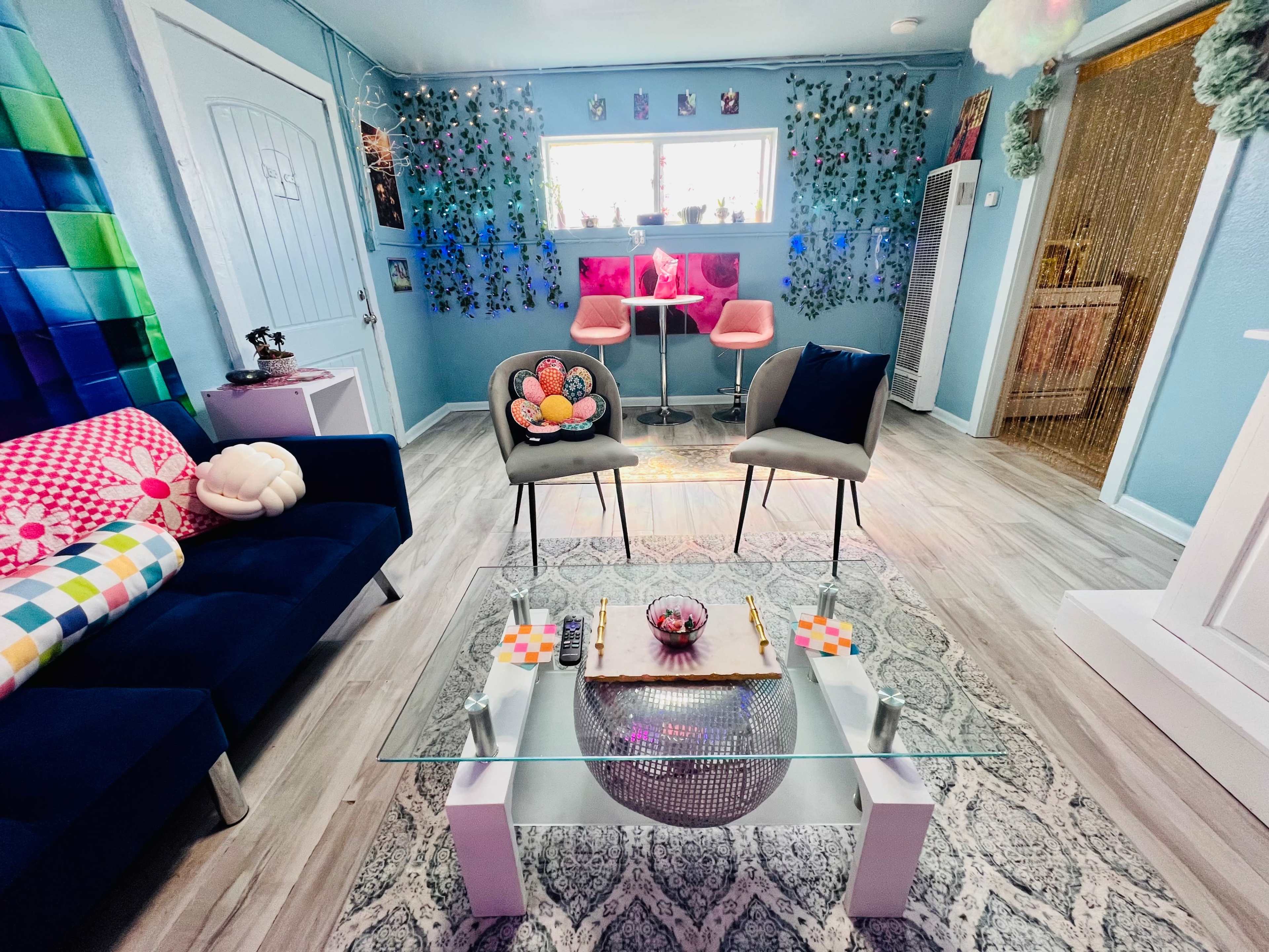 A colorful living room with a glass coffee table, two chairs, a couch adorned with patterned cushions, and decorative plants on the walls.