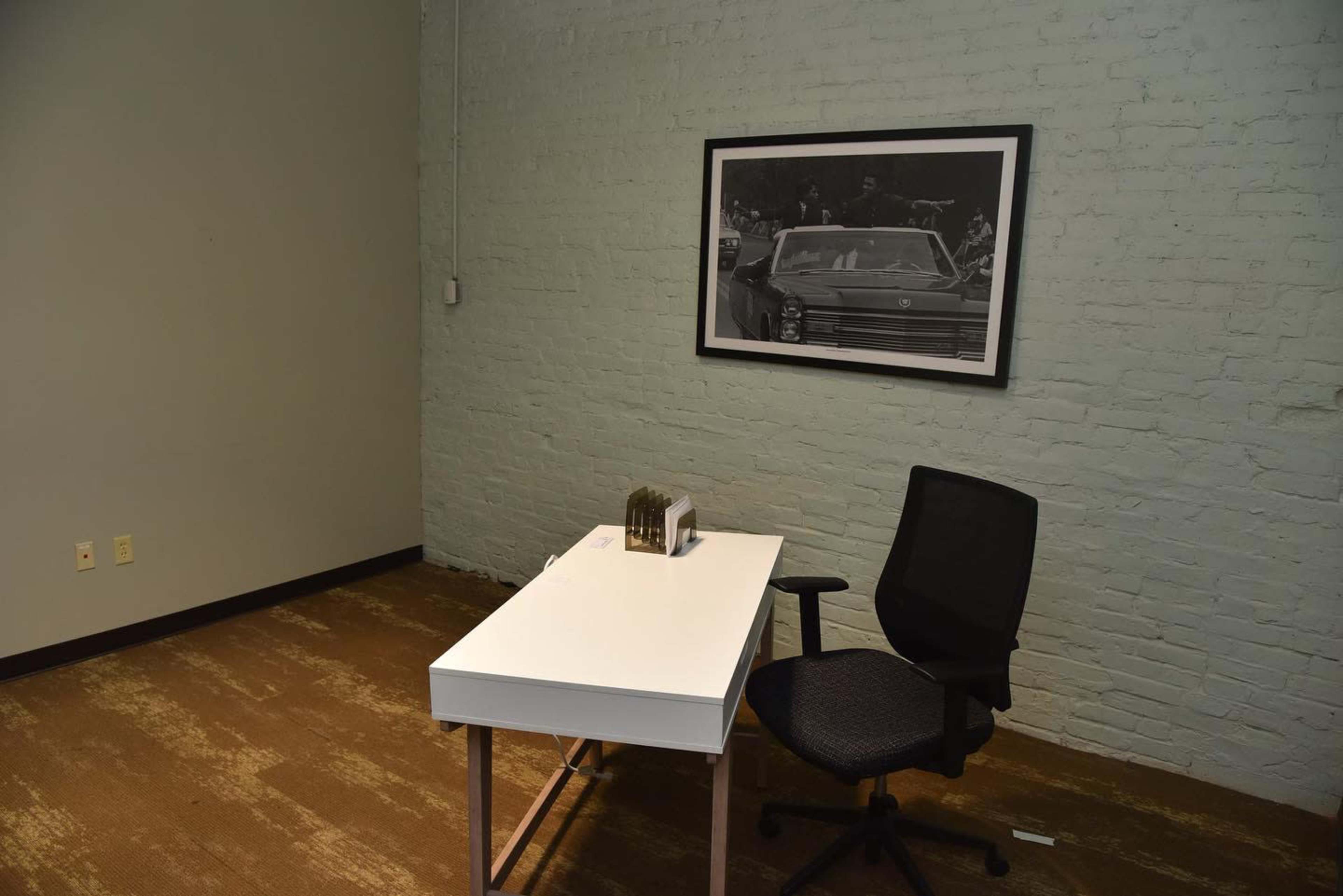A minimalist office space features a white desk and a black chair with a large black-and-white photo of a car on the wall.
