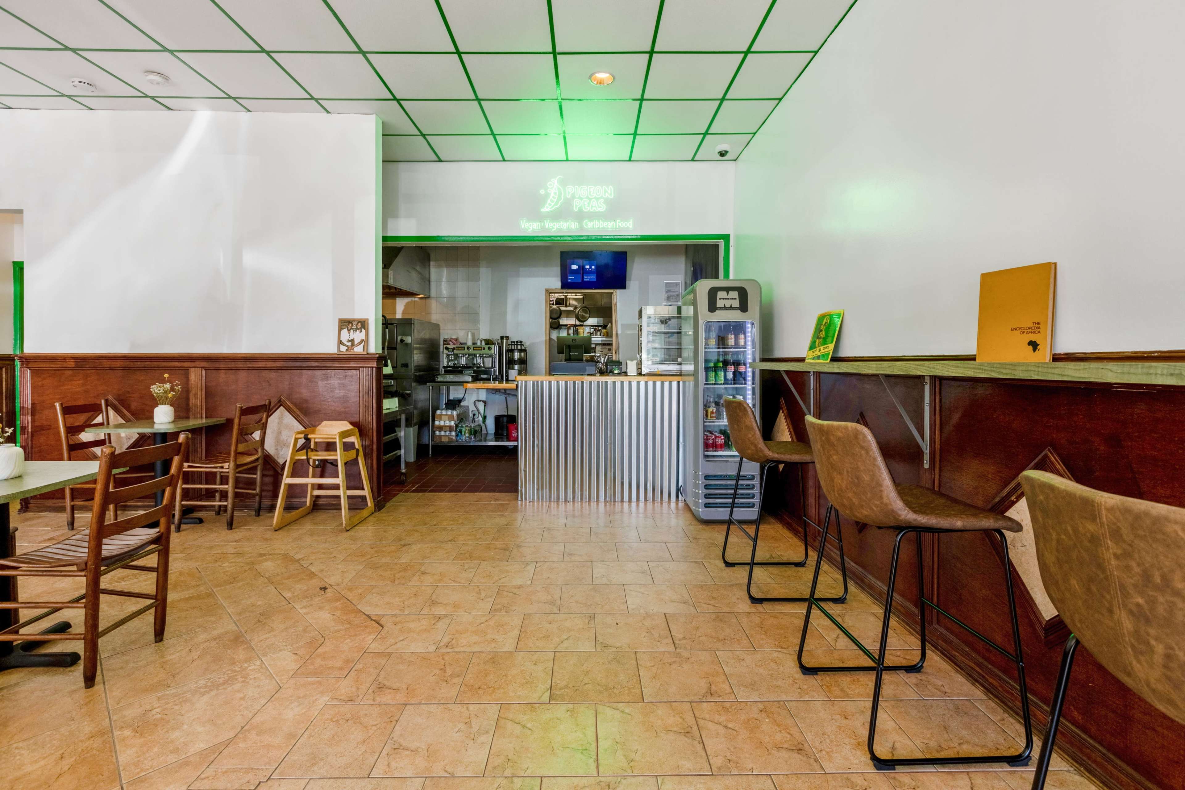 A casual eatery with a service counter in the background, wooden seating, and a small refrigerator.