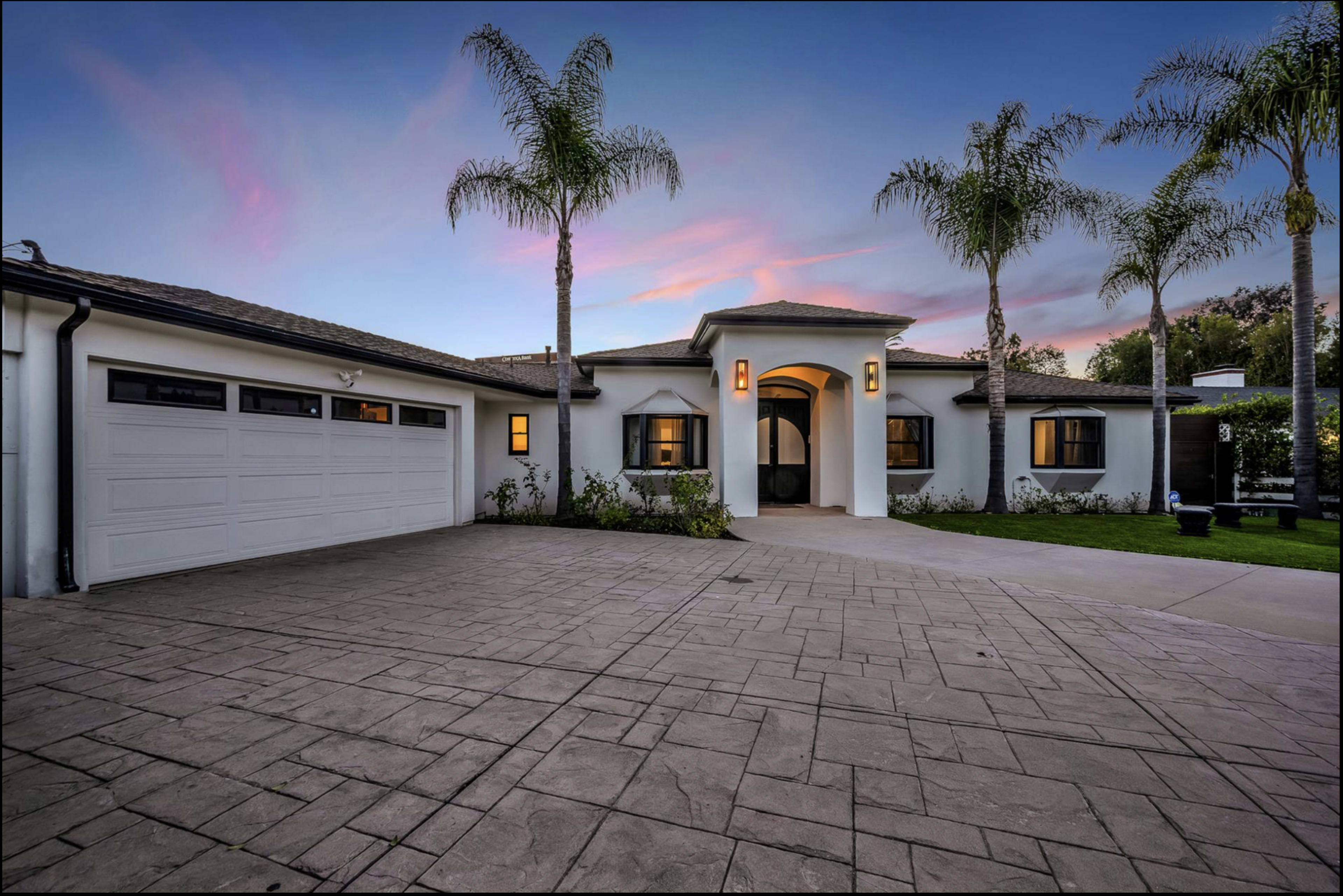 The image shows a modern single-story home with a curved entrance, surrounded by palm trees and a spacious driveway.