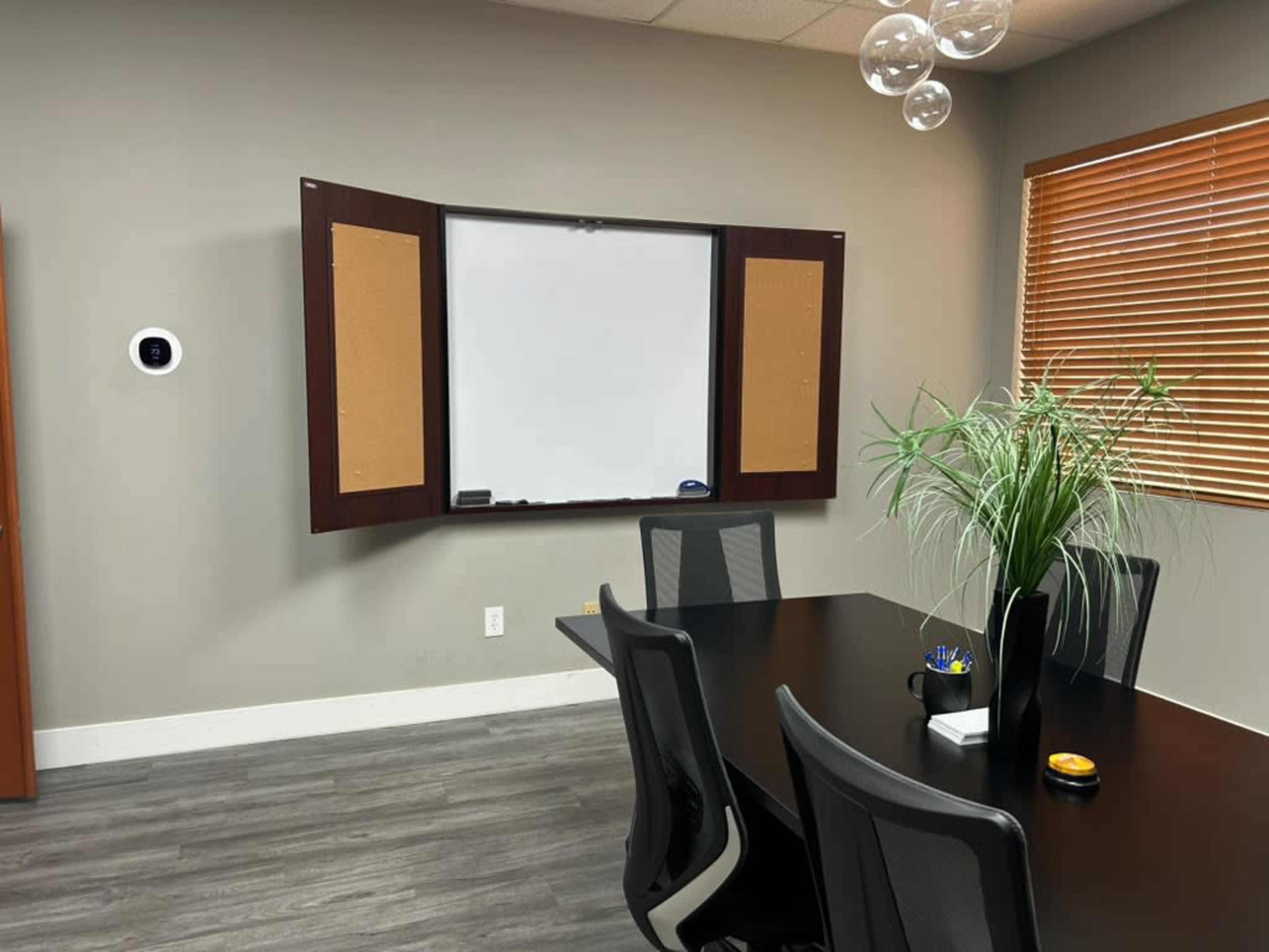 A modern conference room features a black table surrounded by five chairs, with a whiteboard mounted on a wall and a plant in the corner.