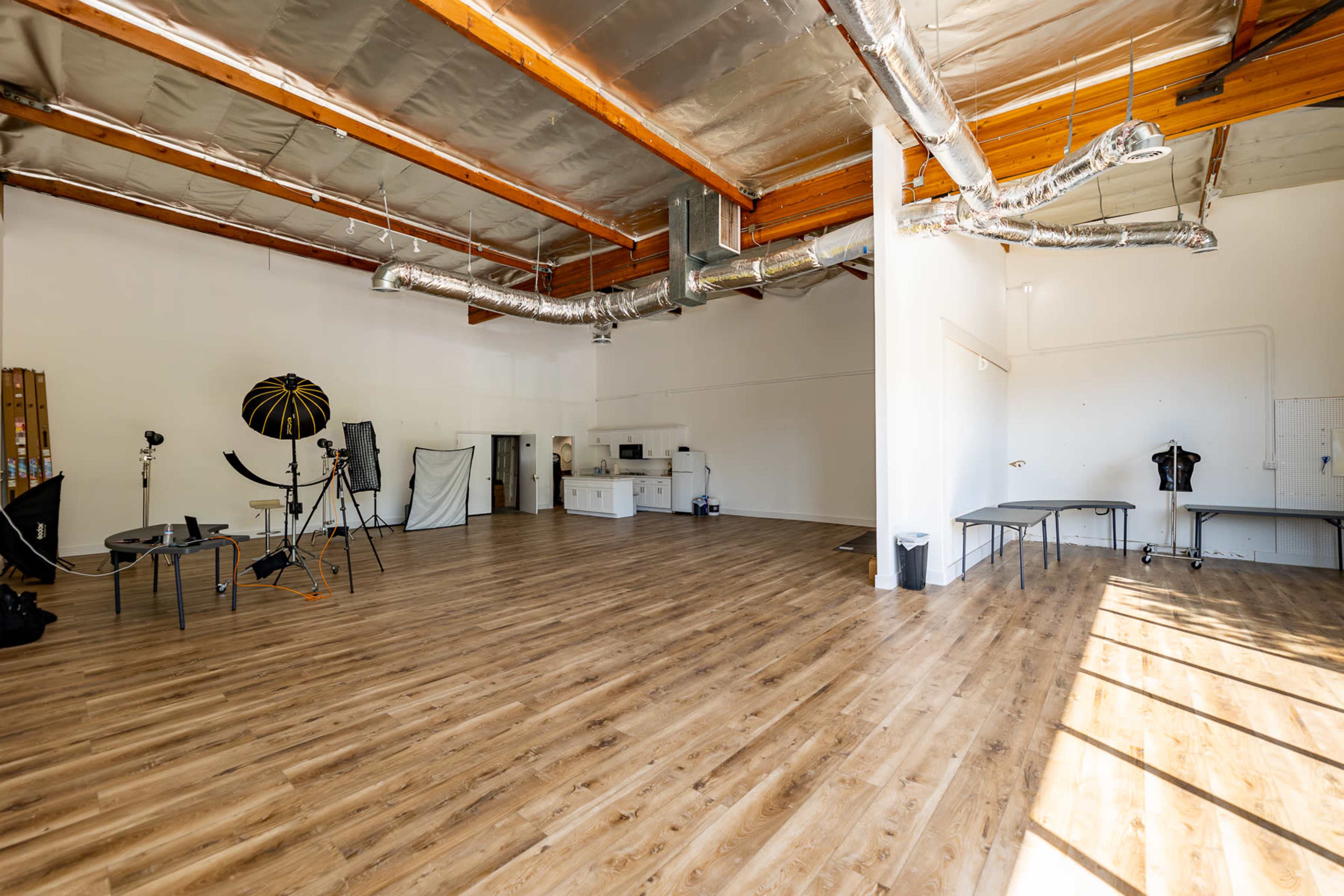 A spacious photography studio with wooden floors, white walls, and various equipment set up, including a stool and tables.