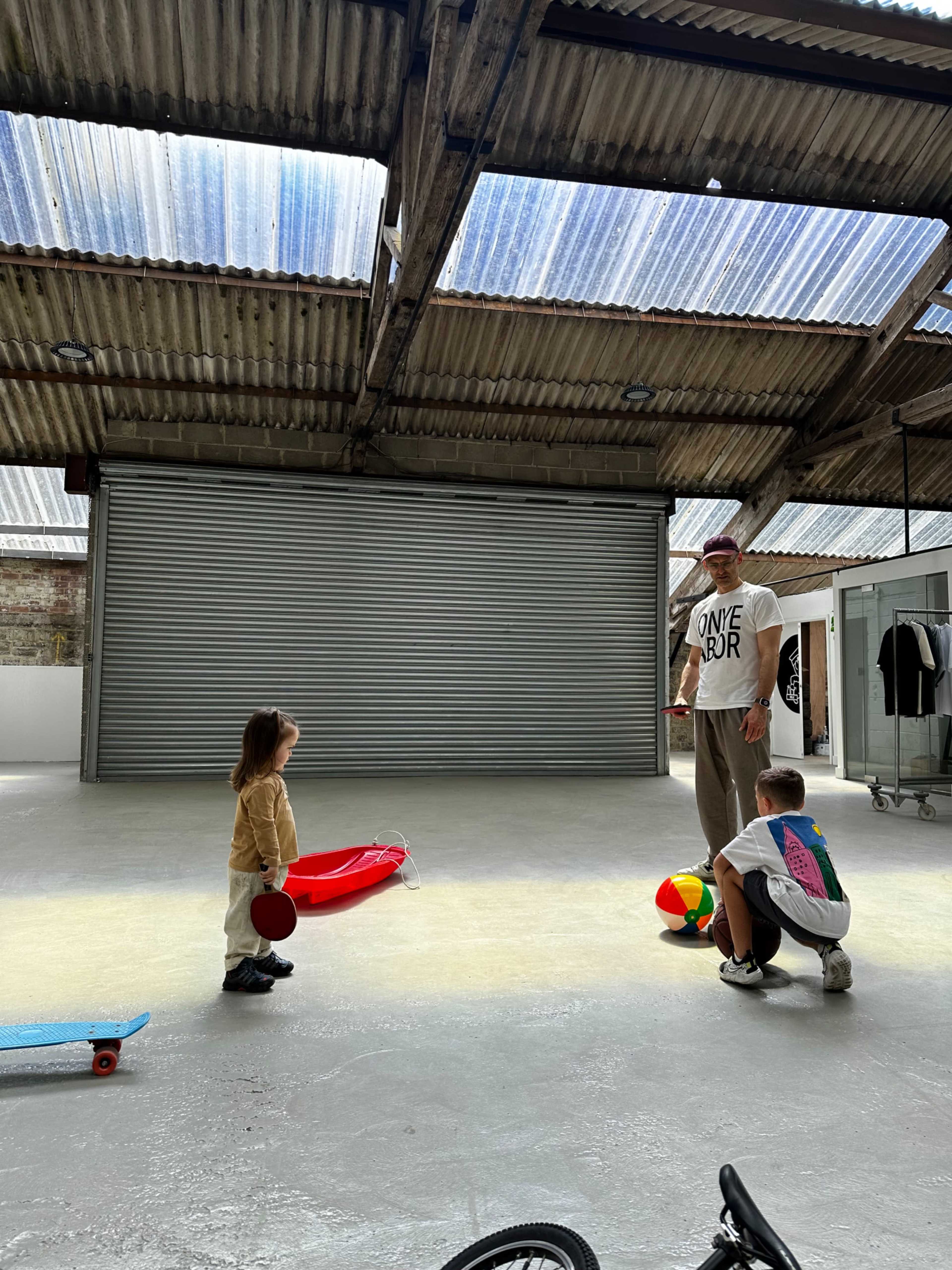 A man stands in a large industrial space with two children, one holding a skateboard and the other a ball, while a red skateboard sits on the floor nearby.