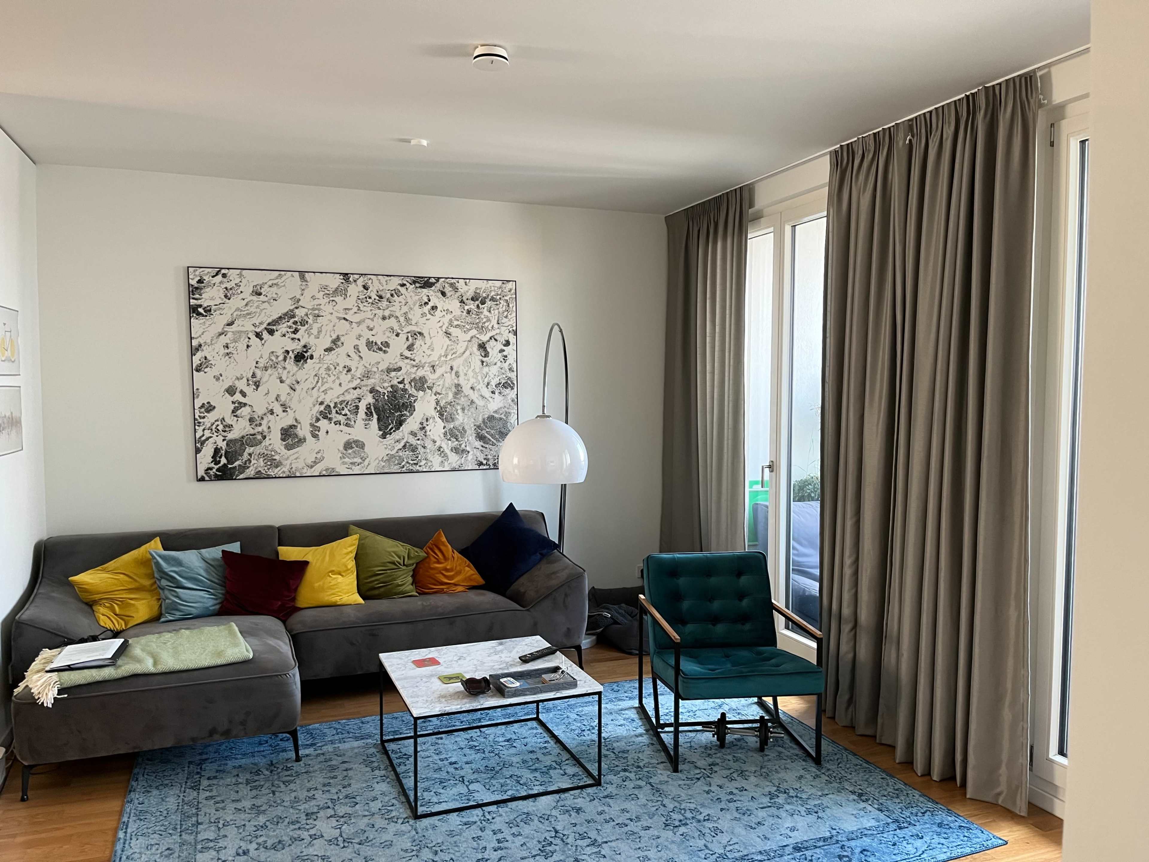 A modern living room features a gray sofa with colorful cushions, a glass coffee table, a green chair, and floor-to-ceiling windows with sheer curtains.