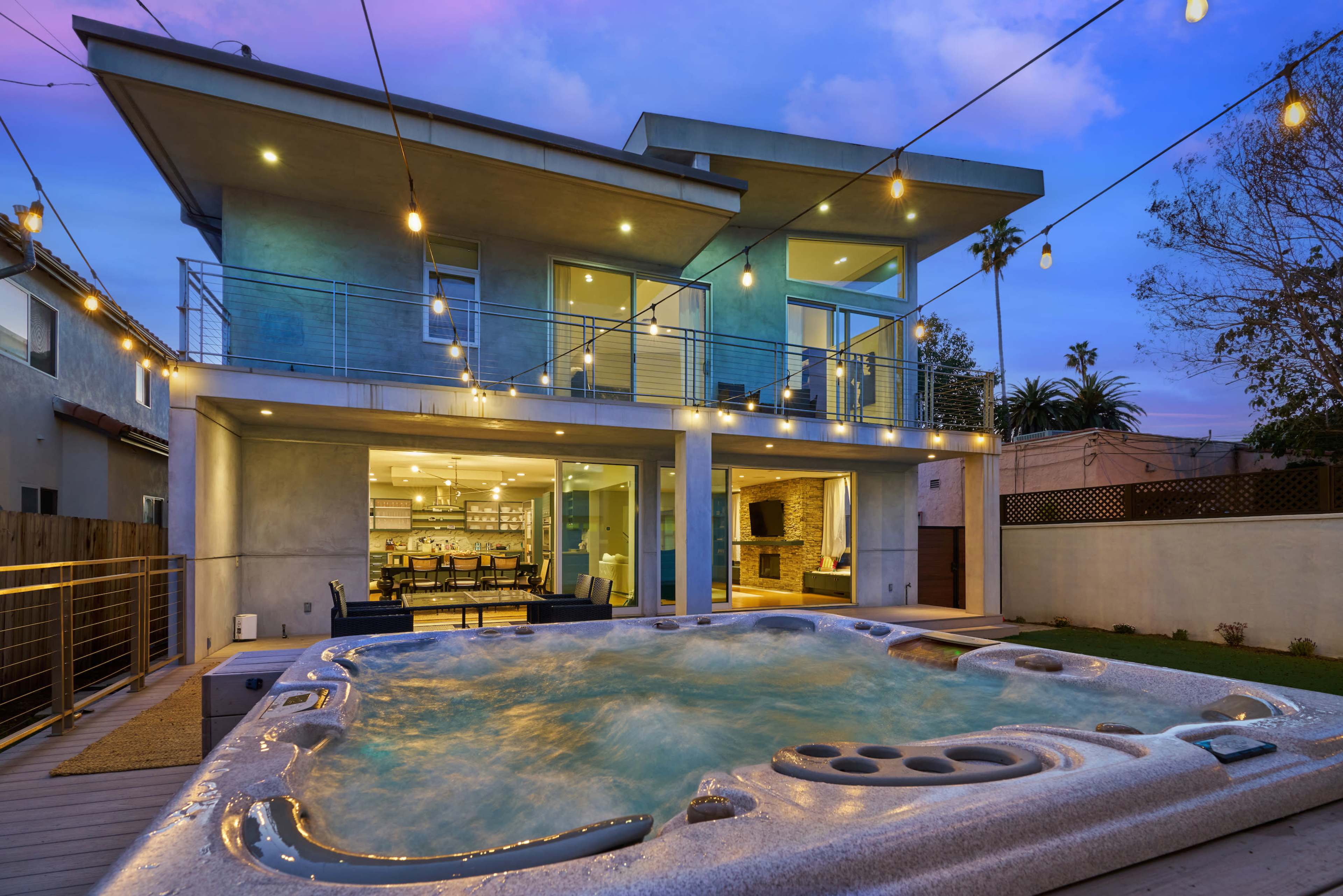 A modern house with a jacuzzi and patio area is illuminated by string lights in the evening.