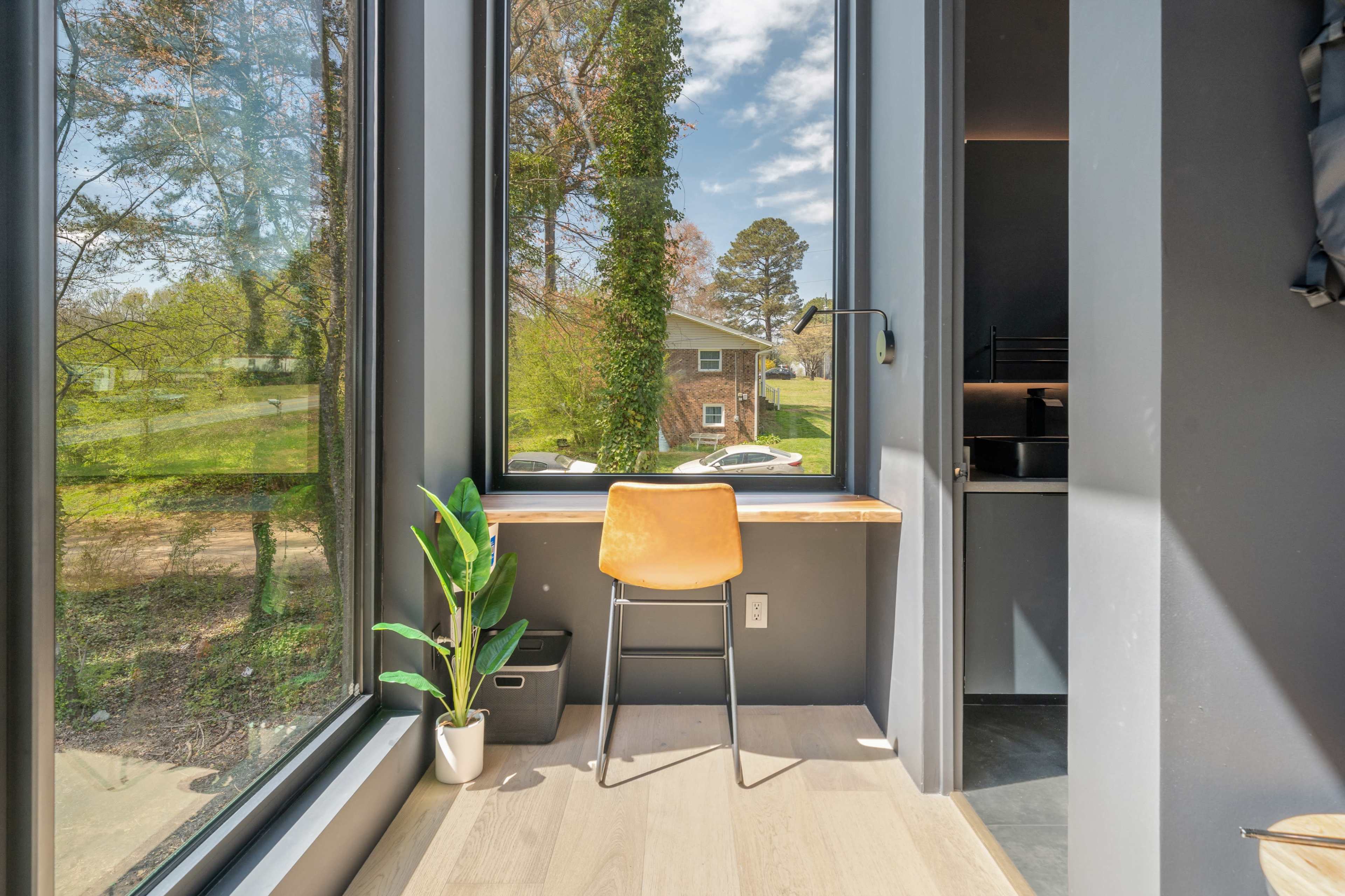 A small workspace with a wooden desk and a chair is set beside a large window, overlooking a green yard and trees.
